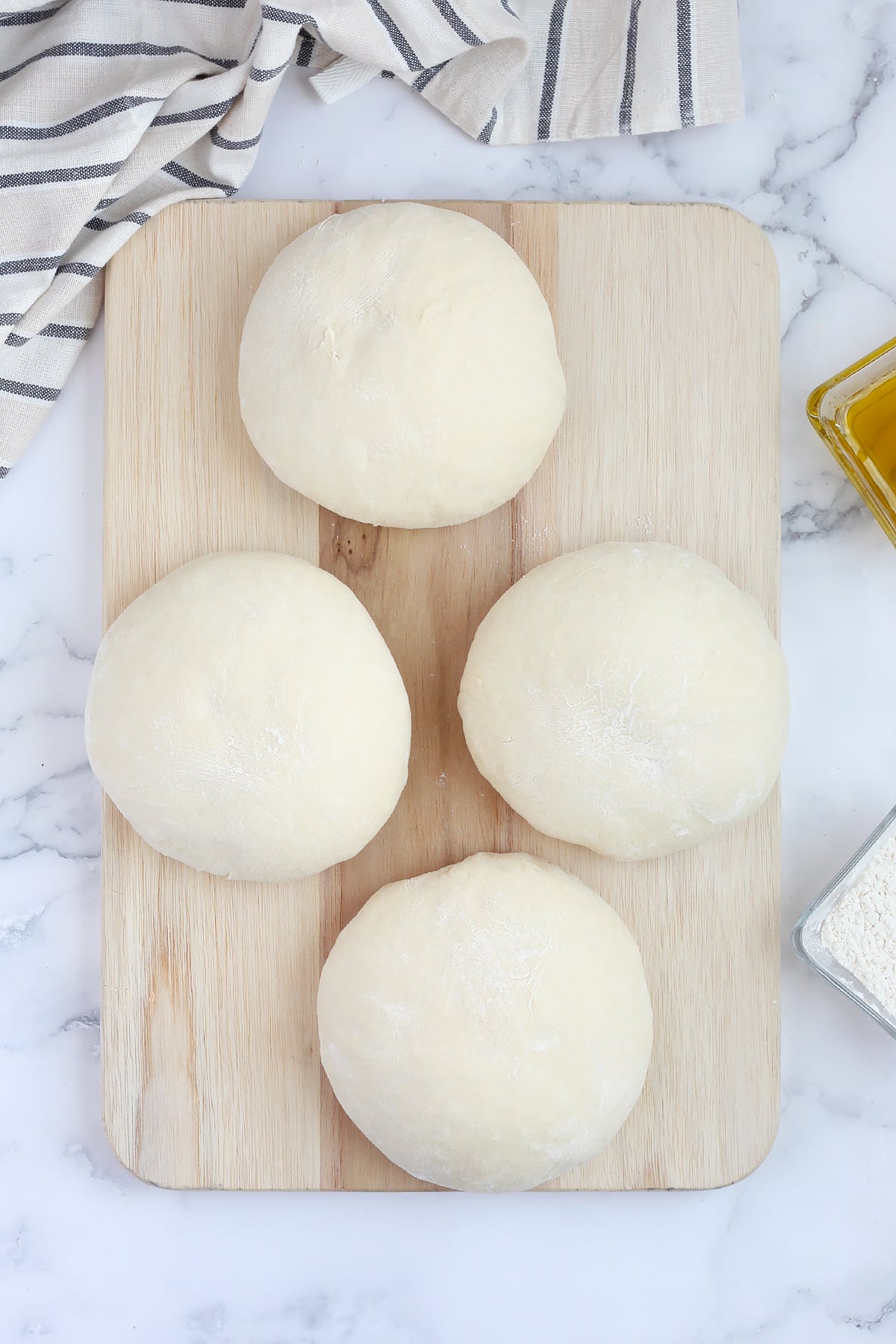 Four balls of pizza dough on a wooden cutting board with a striped linen in the background.