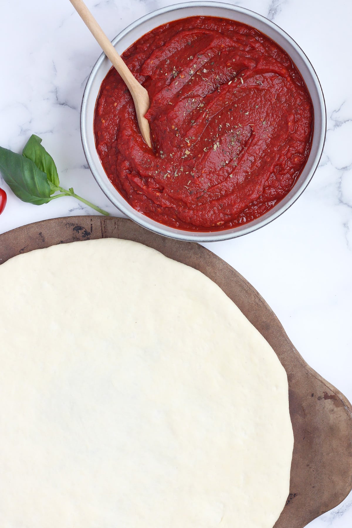 A bowl of pizza sauce near pizza dough rolled out onto a pizza stone.