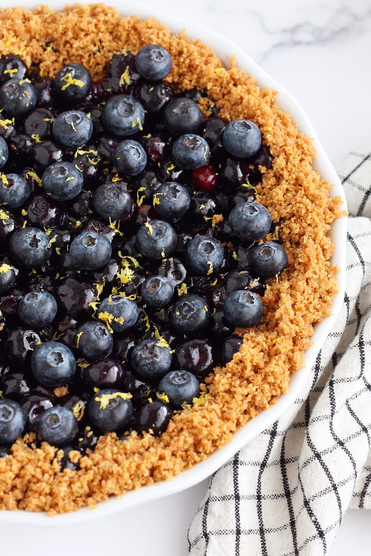 A close-up shot of a blueberry pie with lemon zest and a graham cracker crust crumble.