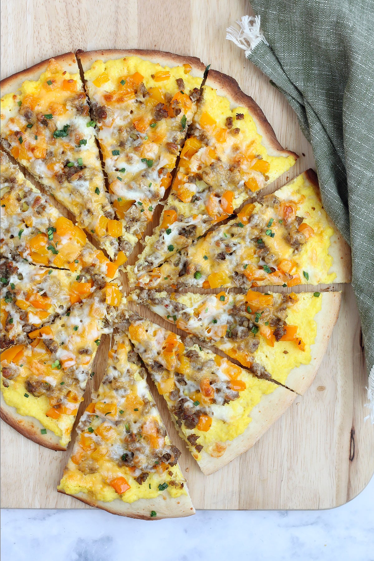 A sliced breakfast pizza topped with sausage, bell peppers and eggs on a wooden cutting board with a green linen.