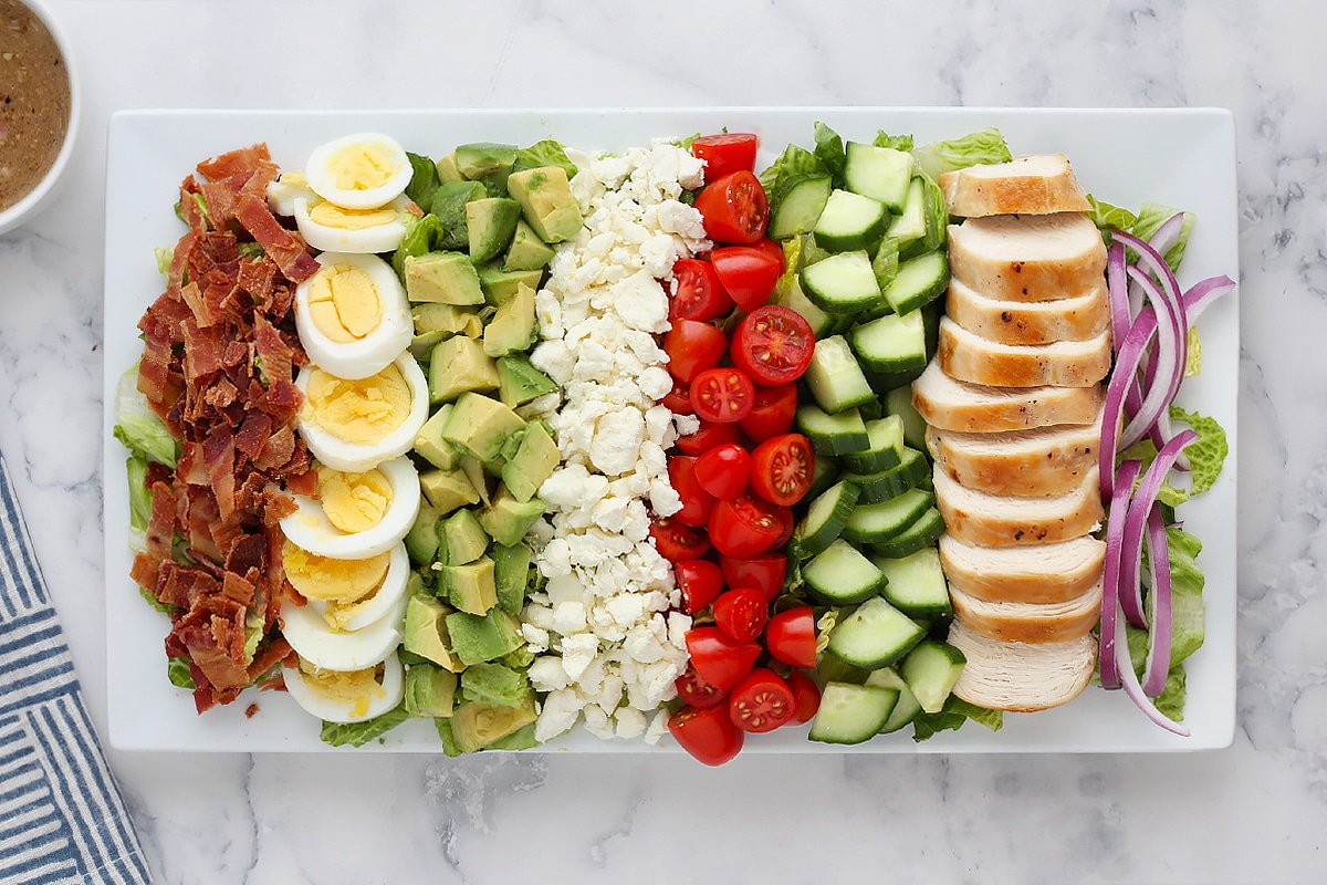 Bacon, eggs, avocado, cheese, tomatoes, cucumbers, chicken and red onion layered on a white serving platter.