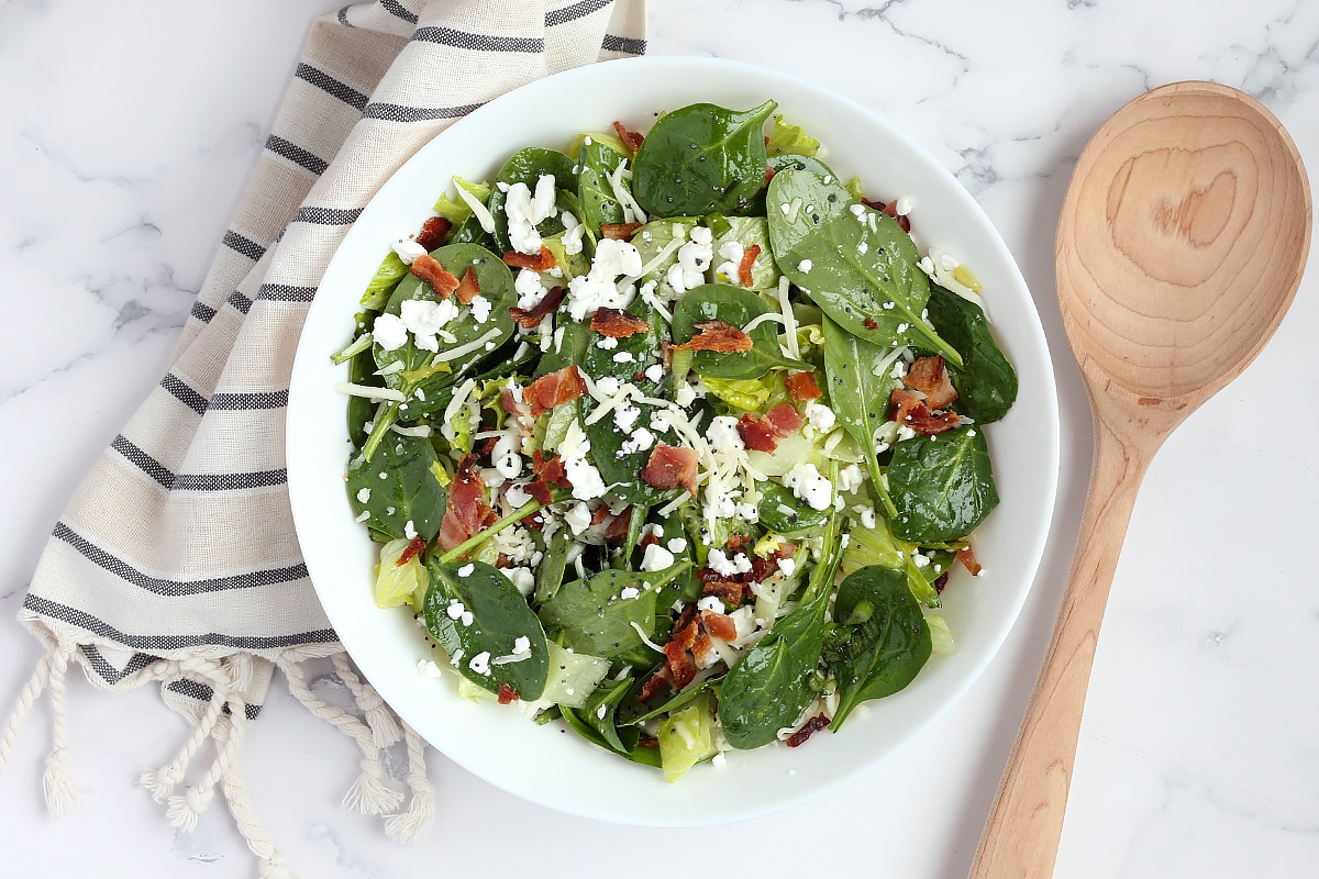 An overhead shot of spinach salad with a striped linen and wooden serving spoon.