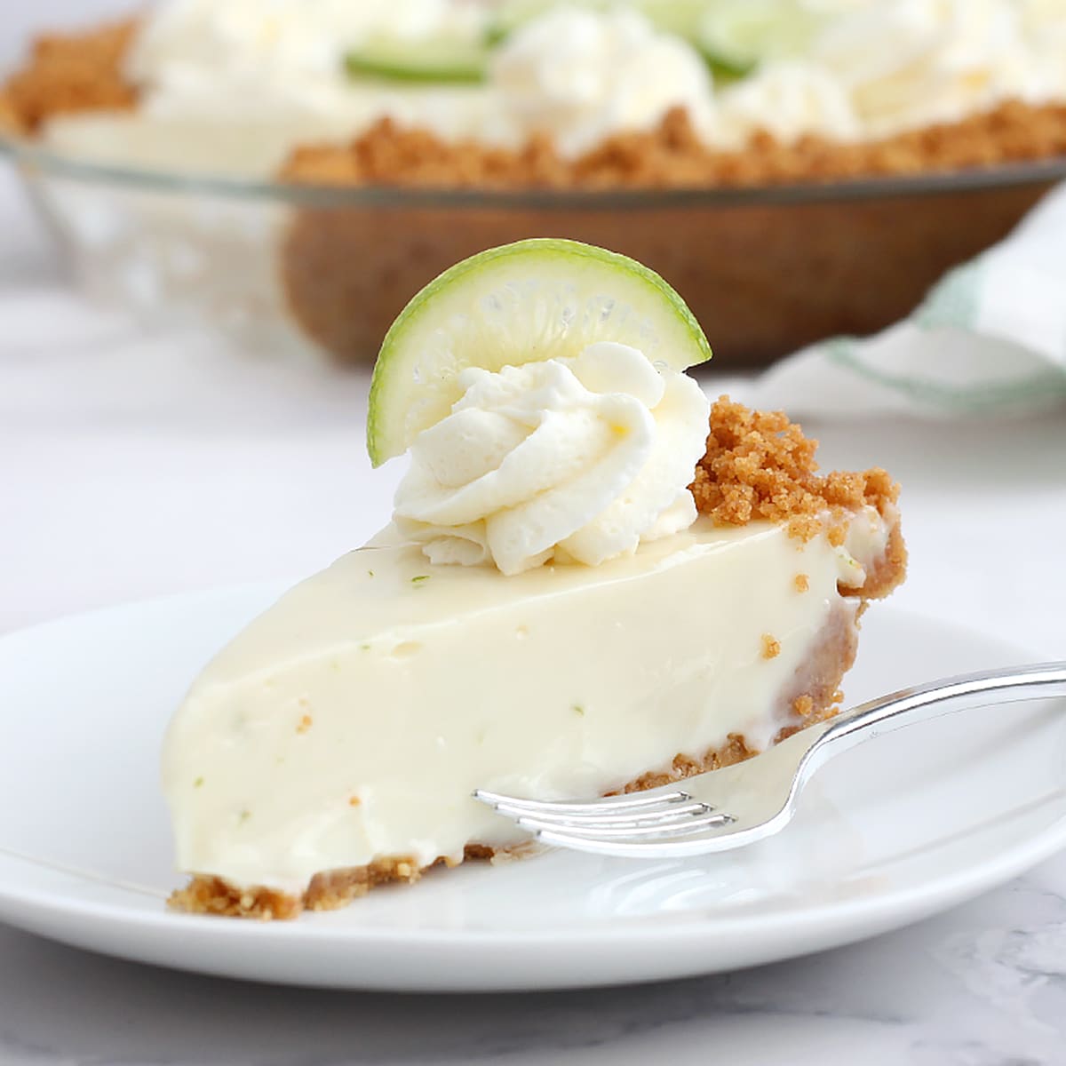 A slice of key lime pie in a graham cracker crust with a swirl of whipped cream and a slice of lime.