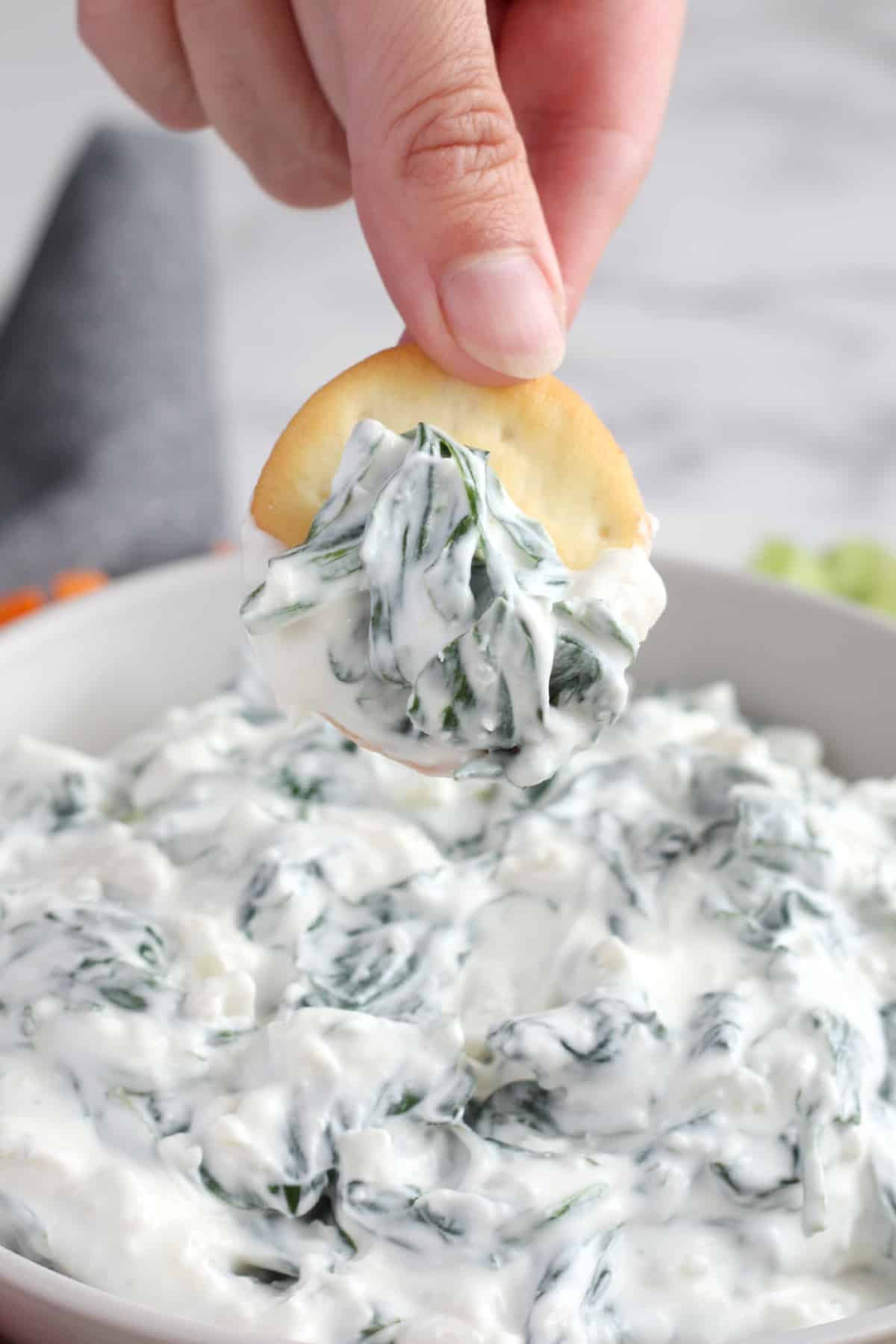 Someone scooping spinach dip out of a bowl with a cracker.