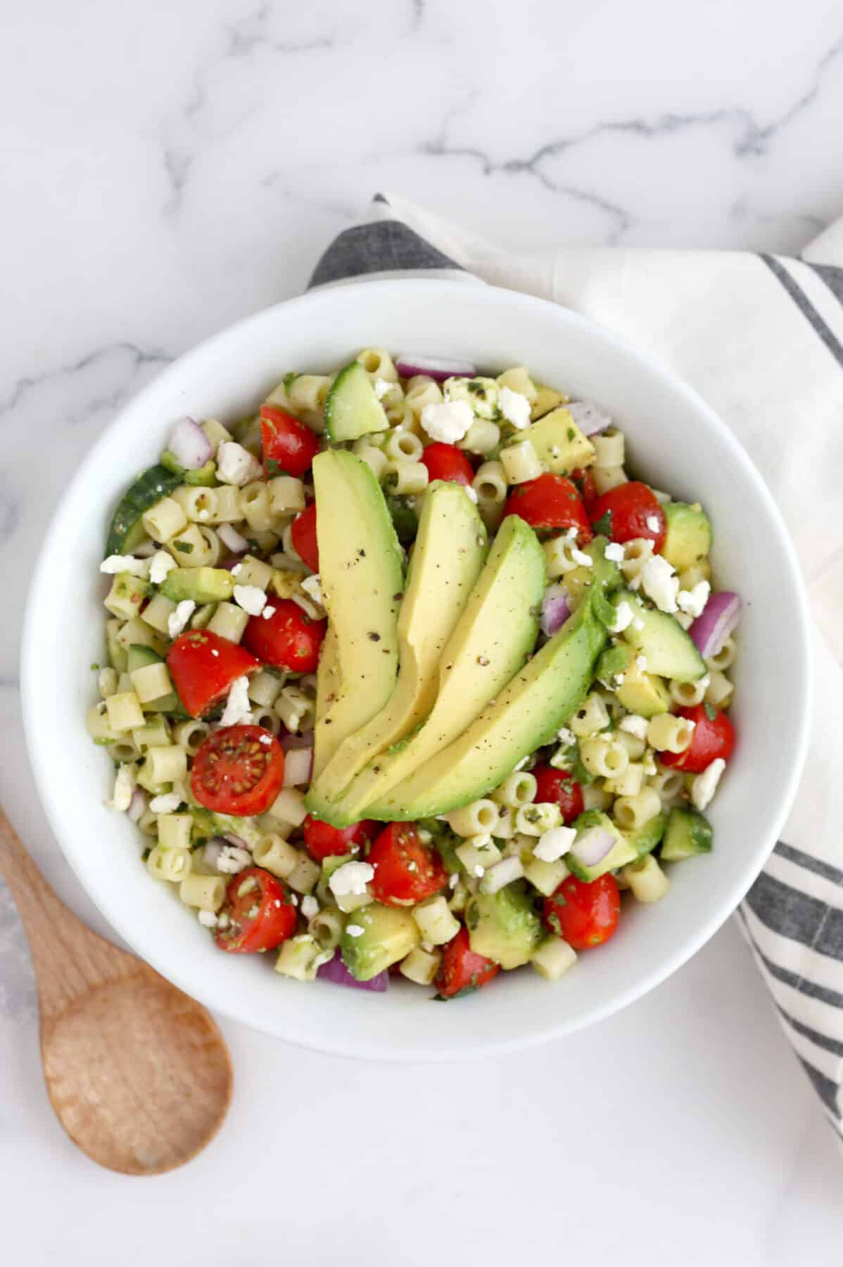 Avocado salad with pasta and veggies tossed with a cilantro lime dressing in a white bowl with a striped linen. 