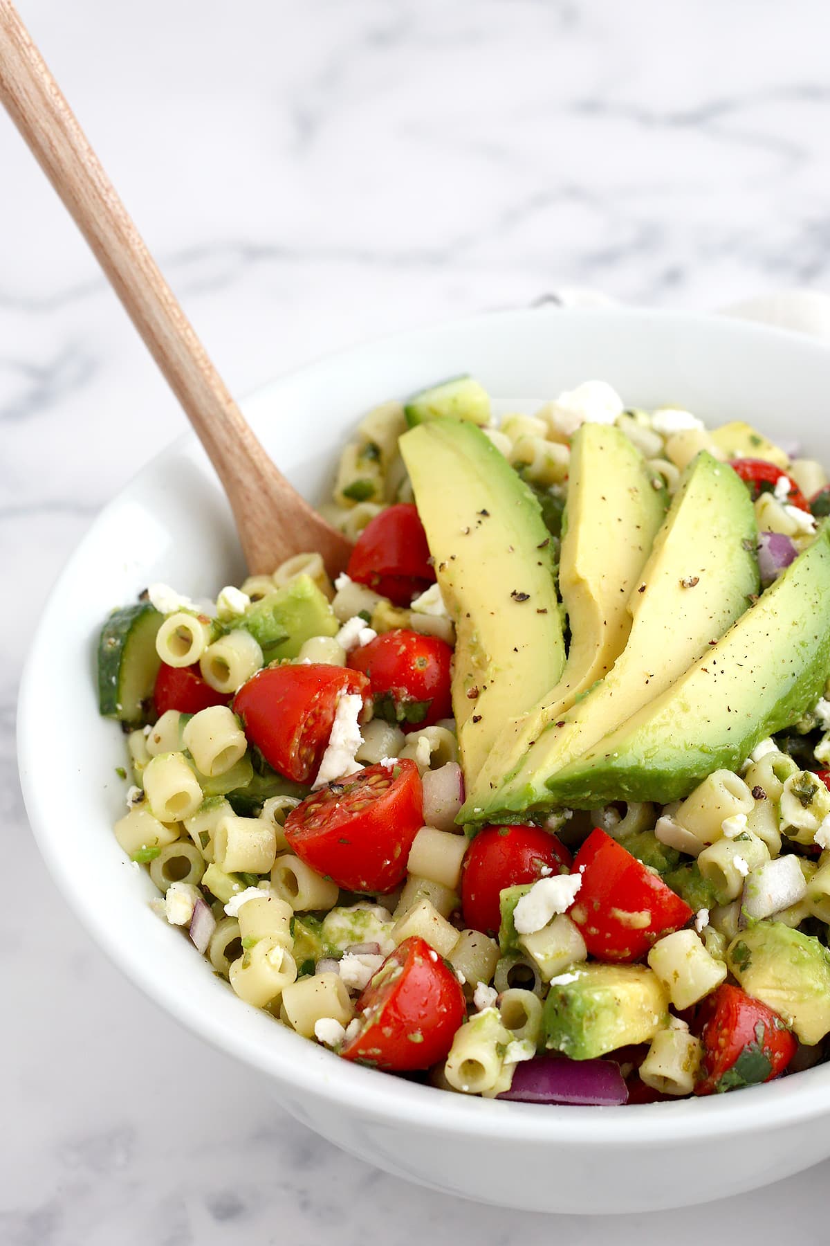 Pasta, tomatoes, cucumbers and avocado in a bowl with a wooden serving spoon.