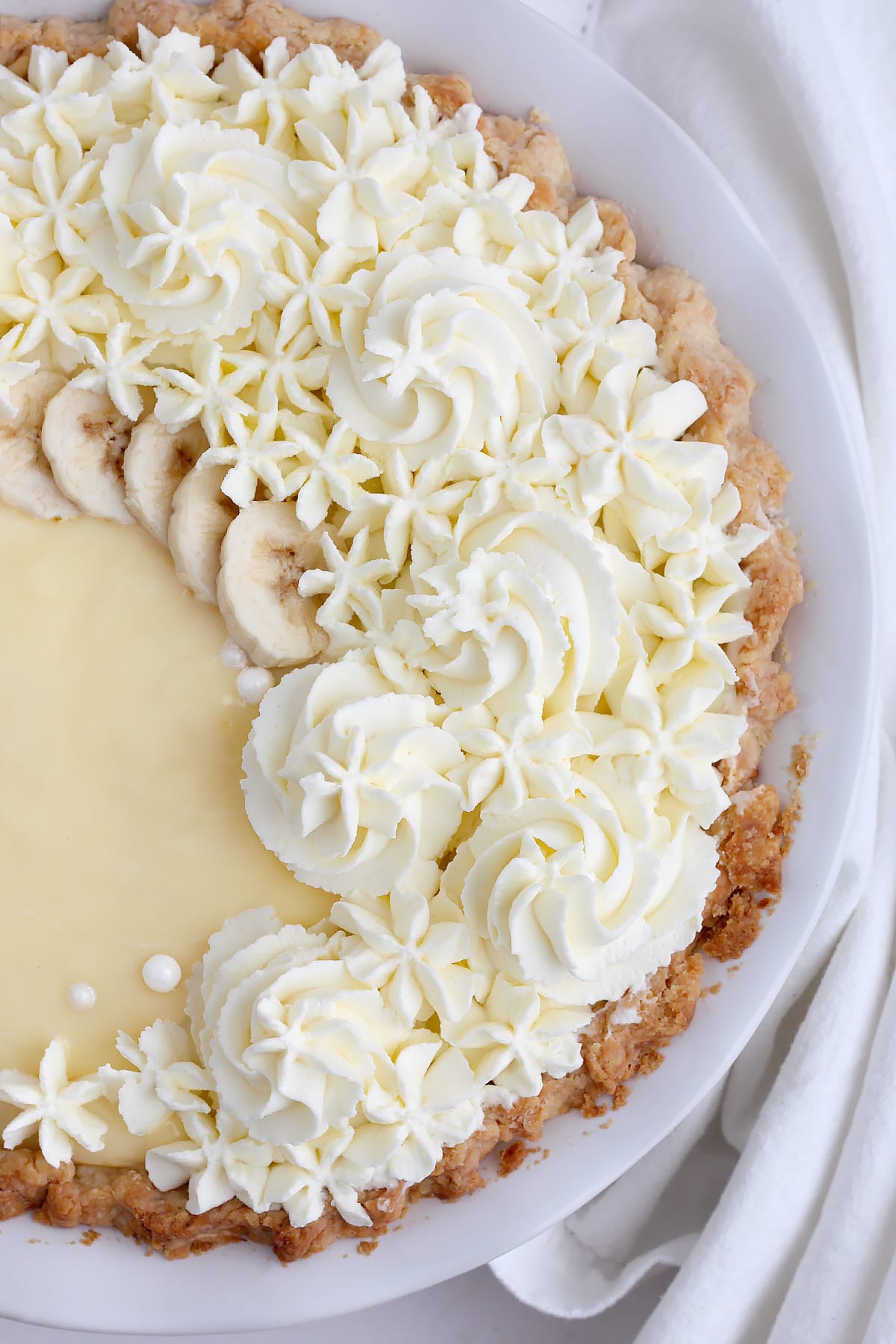 A close up photo of banana cream pie topped with fresh banana slices, whipped cream decorations and candy pearls.