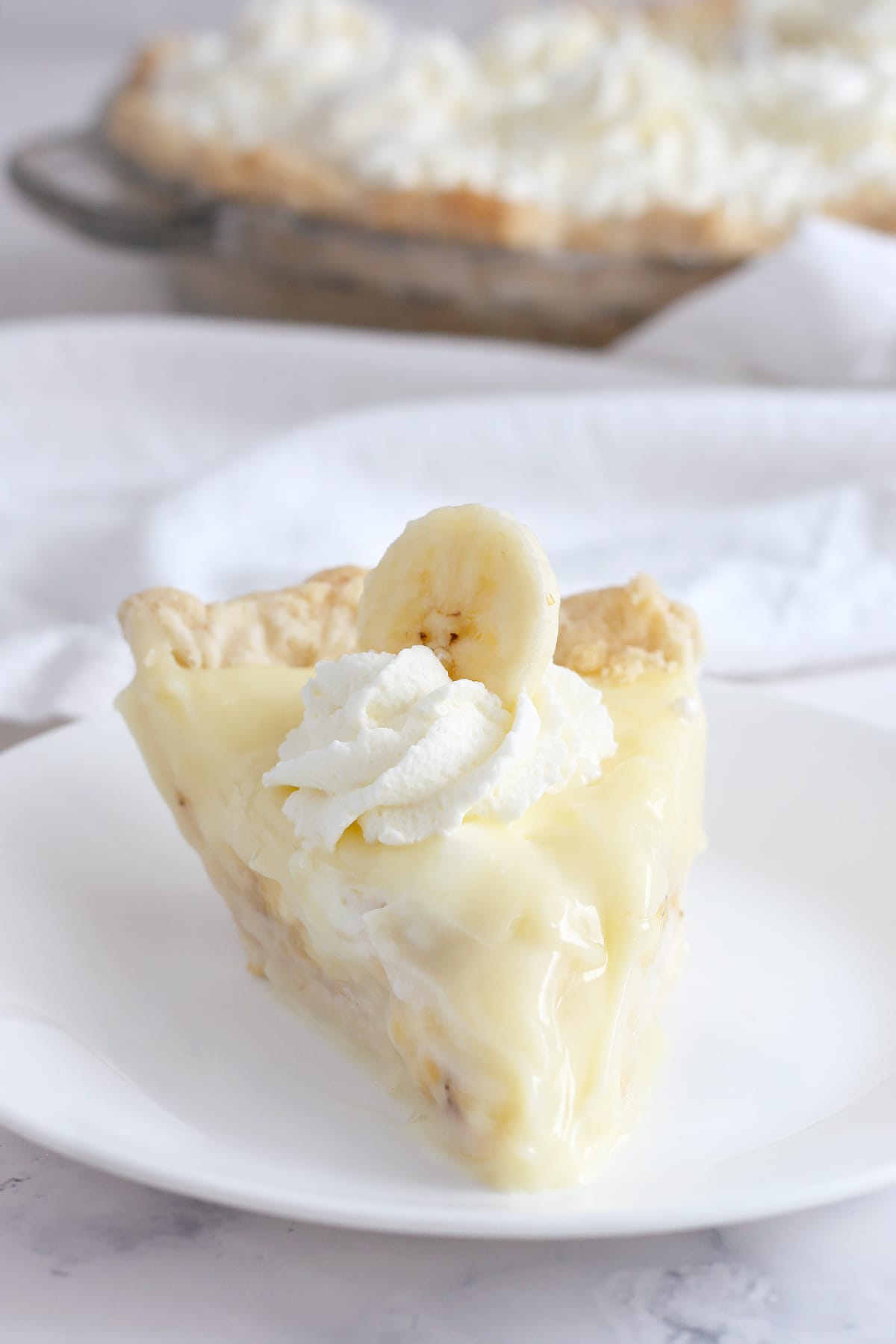 A slice of banana cream pie topped with fresh whipped cream and a slice of banana on a white serving plate.