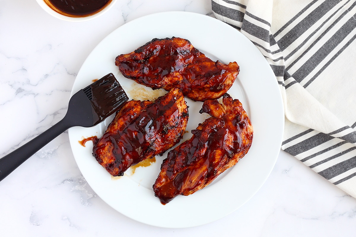 Grilled BBQ Chicken Breasts on a white plate with a pastry brush and a striped linen in the background.