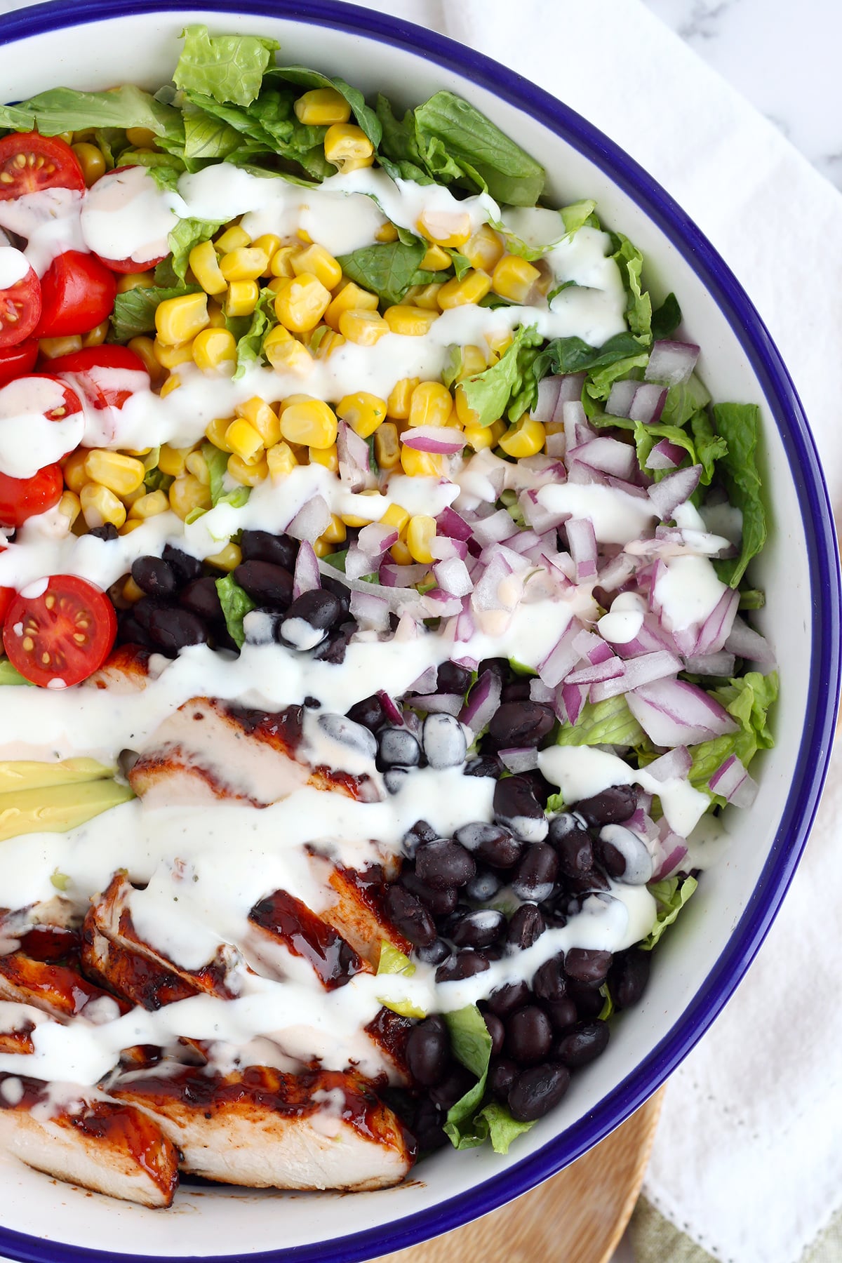 BBQ Chicken Salad with grilled chicken, black beans, avocado, tomatoes and drizzled with a BBQ ranch dressing in a white and blue serving bowl.