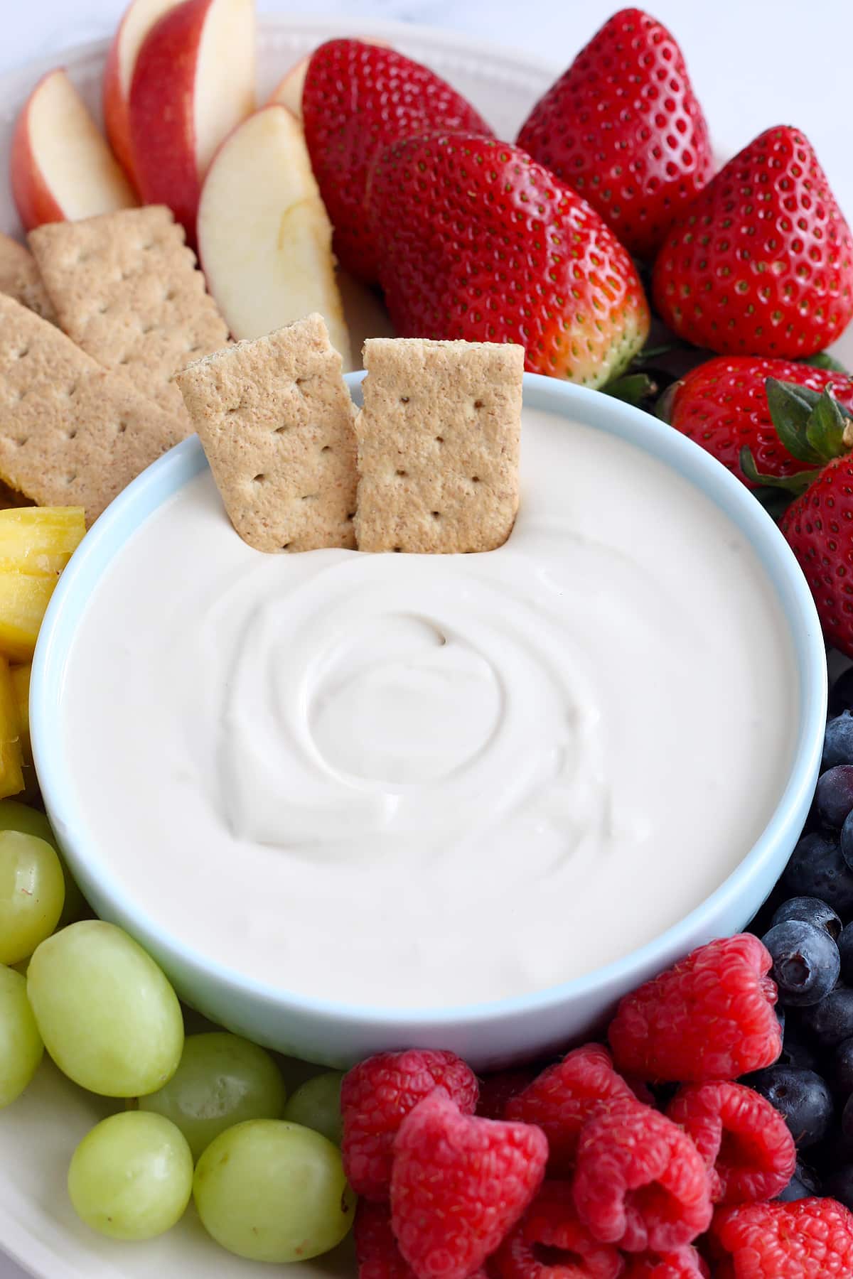 A bowl of creamy fruit dip with strawberries, graham crackers, sliced apples and pineapple.