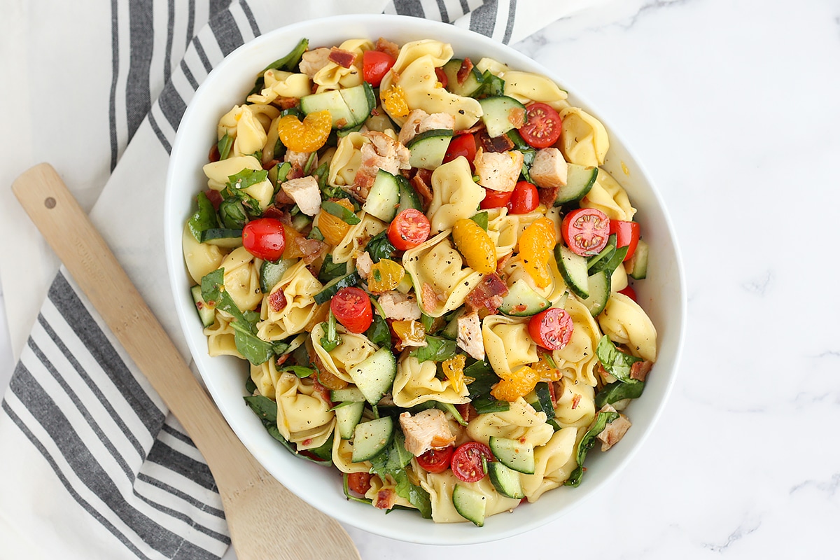 Pasta salad in a large serving bowl with a striped linen and a wooden spoon.