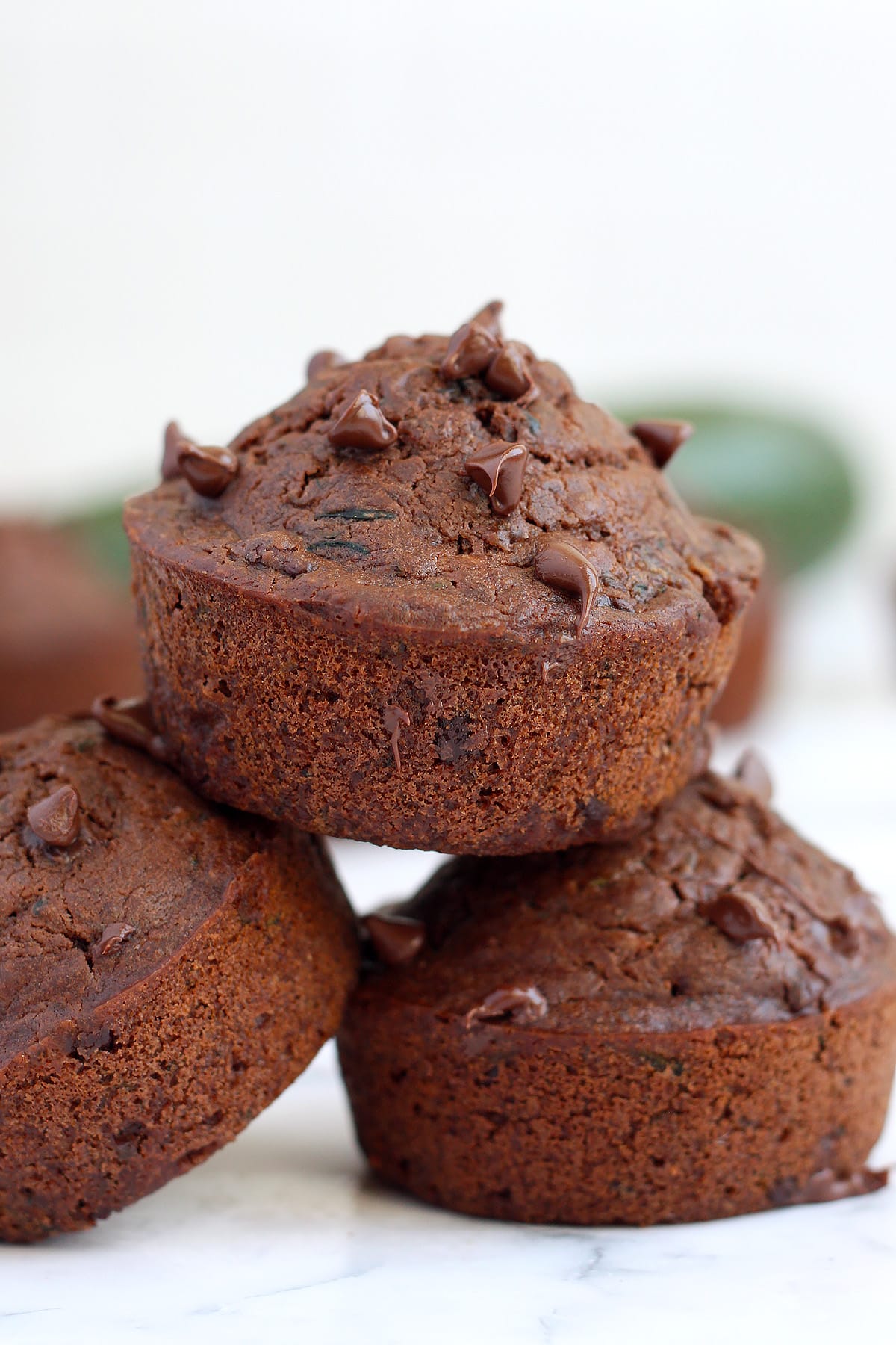 Three double chocolate zucchini muffins stacked on top of each other in front of a zucchini.