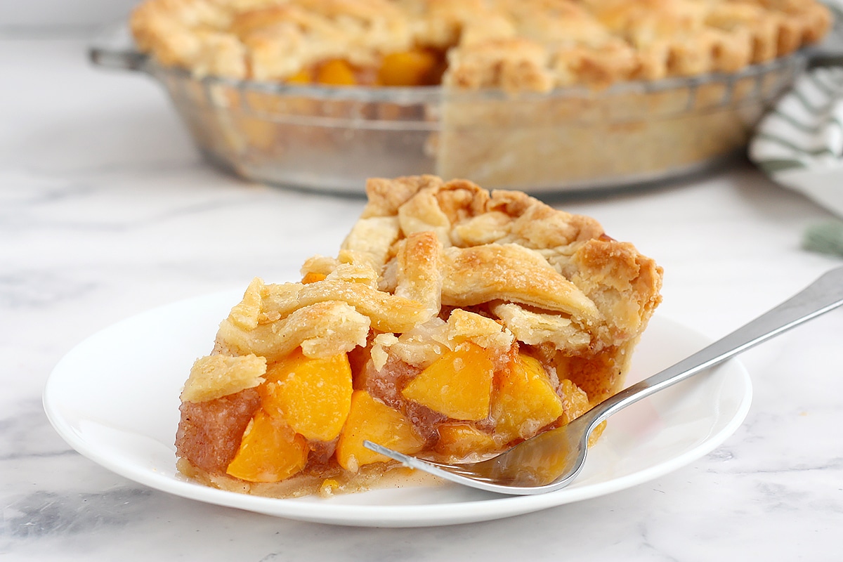 A close up picture of a slice of peach pie on a white plate with a fork.