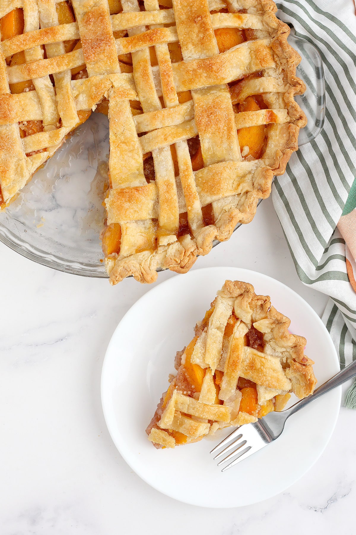 Homemade peach pie with a slice cut out on a white serving plate with striped peach linen in the background.