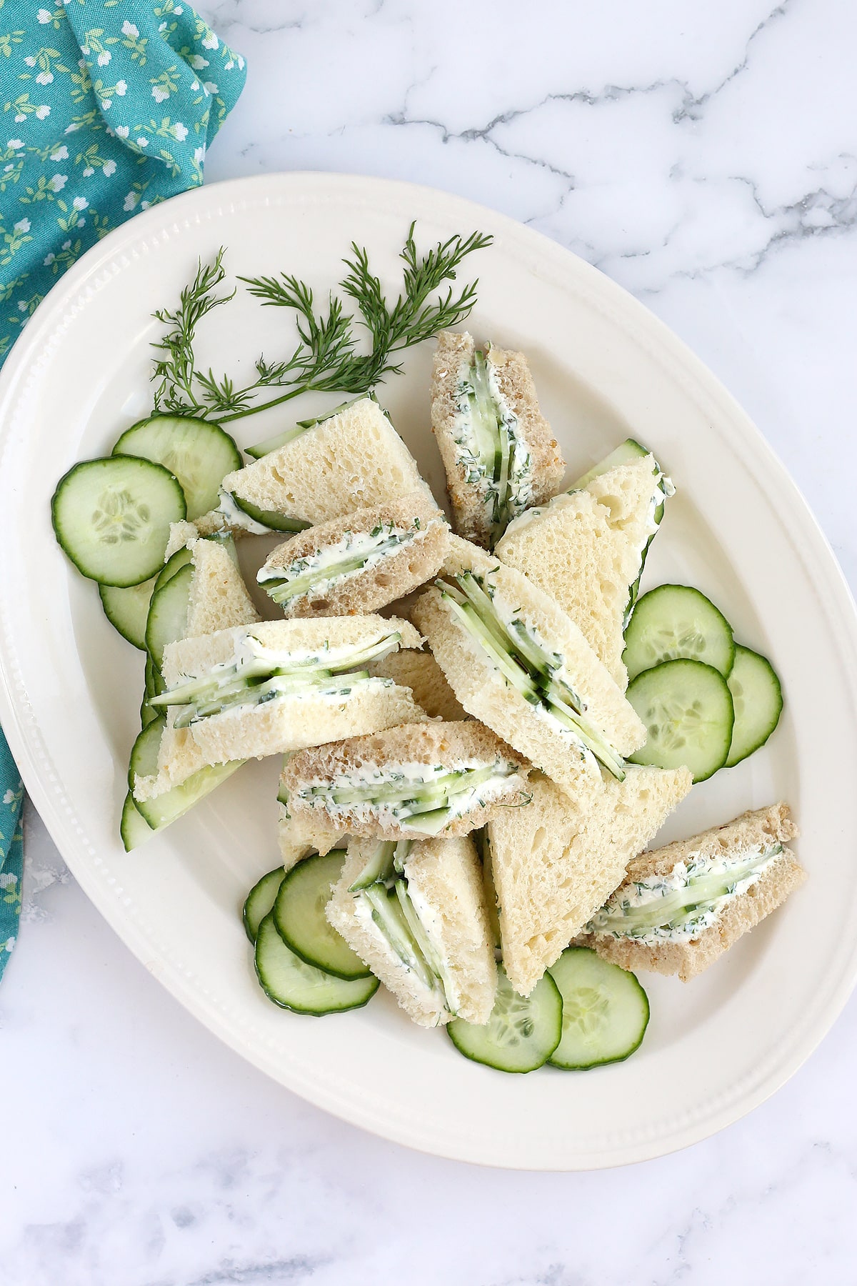 An oval plate topped with cucumber sandwiches, cut into triangles and garnished with fresh dill.