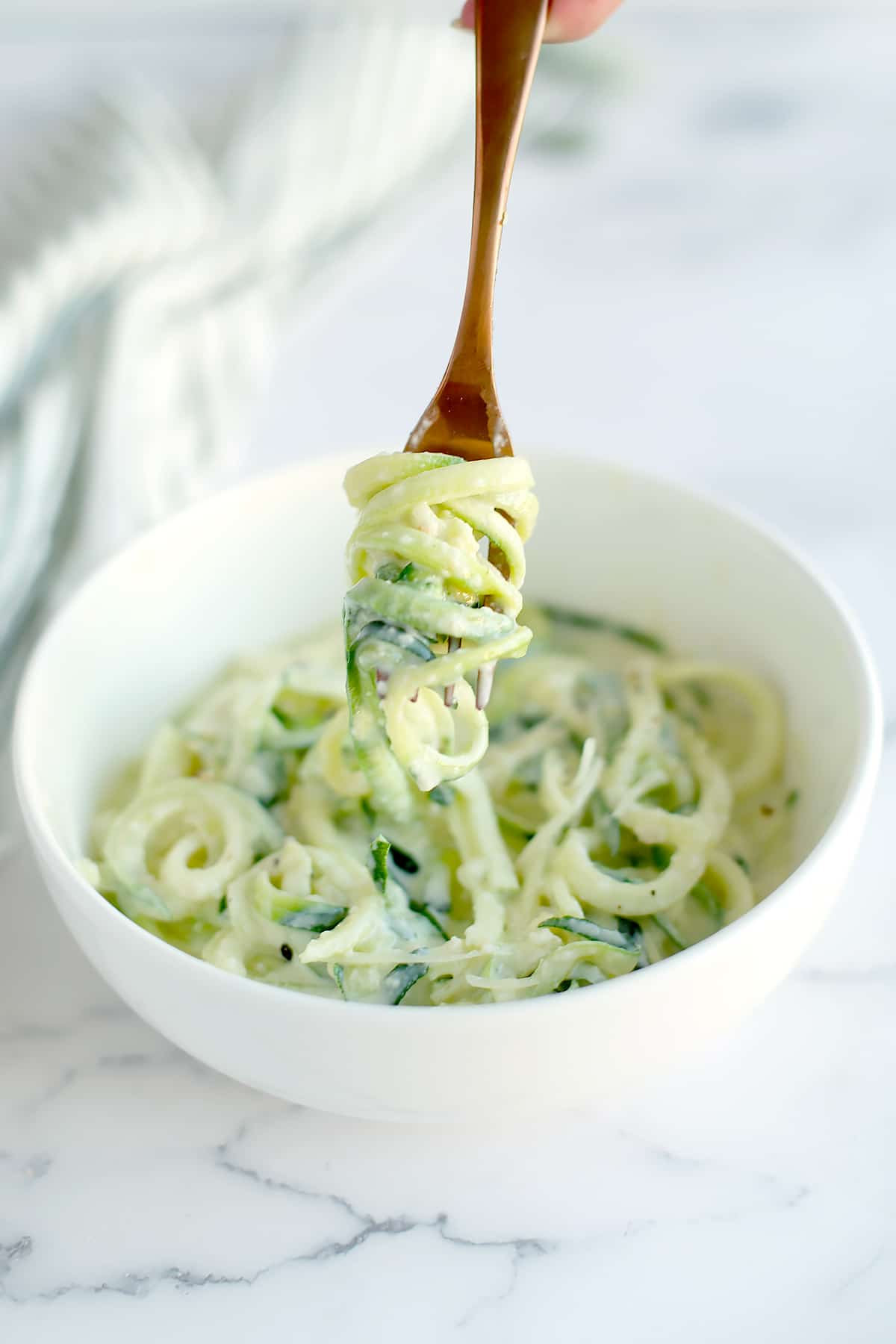 Zucchini noodles in homemade alfredo sauce in a white bowl with a copper fork.