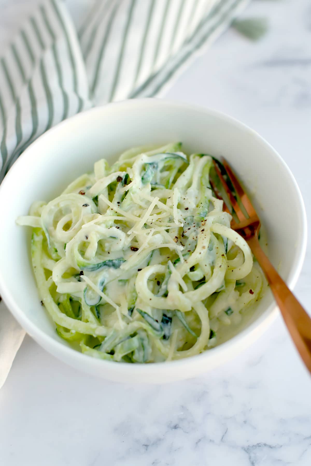 A bowl of creamy alfredo zucchini noodles topped with parmesan cheese and black pepper with a copper fork.