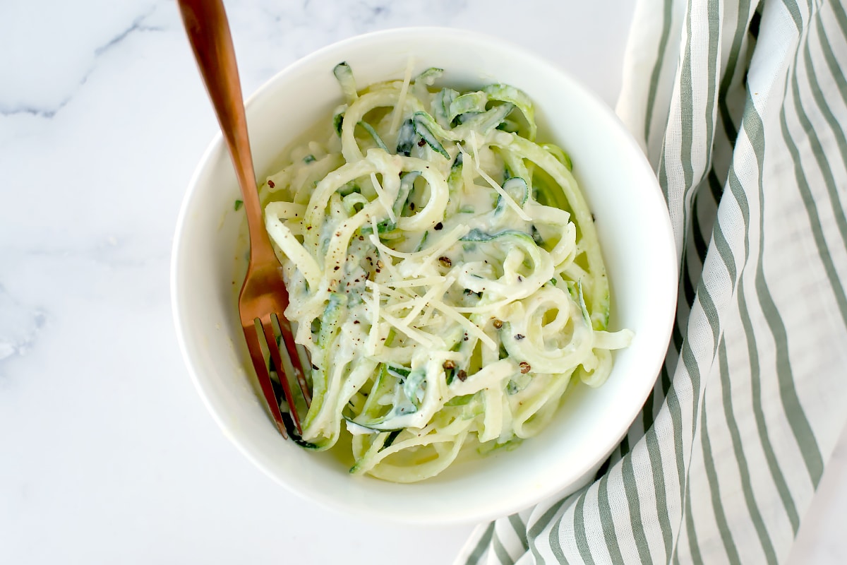 A bowl of zucchini alfredo noodles in a white porcelain bowl with a green striped linen in the background.