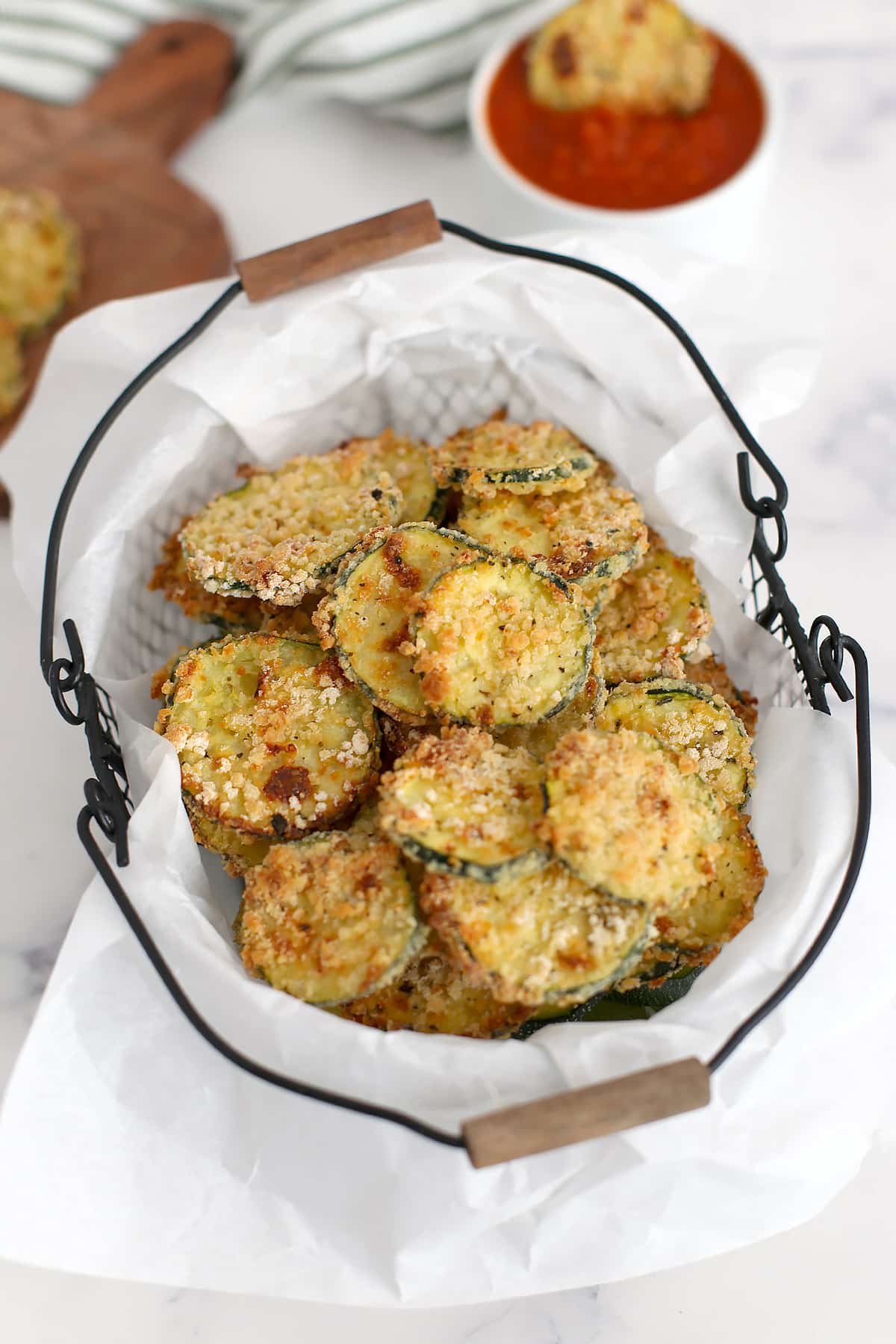 A parchment lined basket filled with crispy baked zucchini chips with a side of marinara sauce in a white bowl.