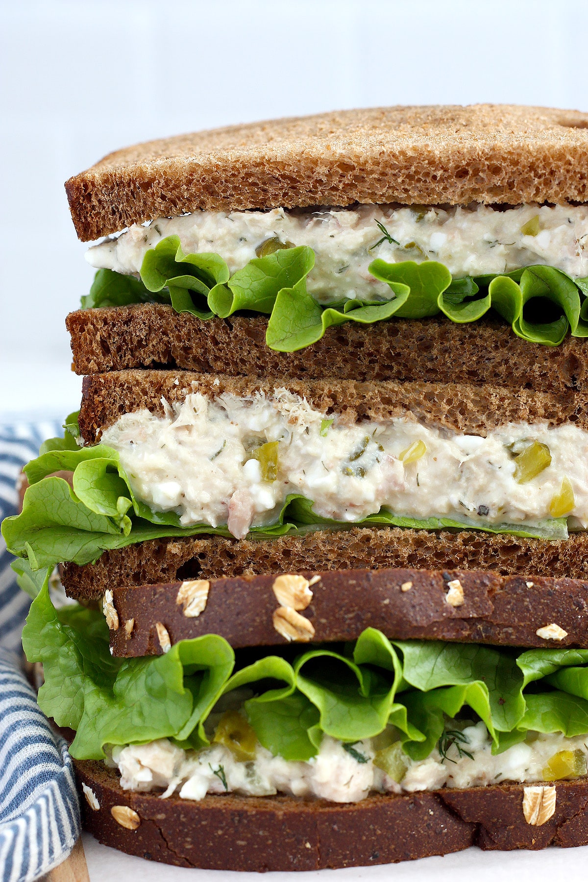 A close up picture of a tuna sandwich with lettuce on rye bread with a blue striped linen in the background.