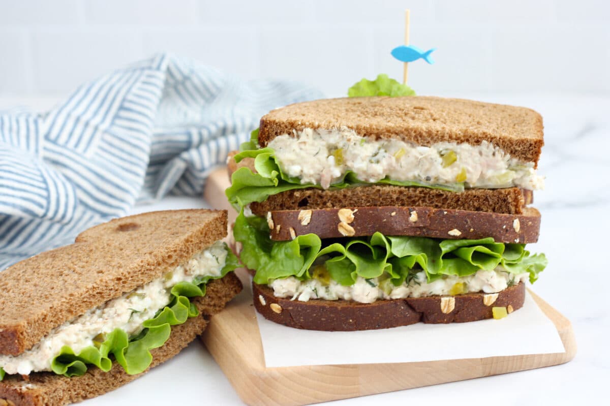A tuna sandwich with lettuce on wheat bread stacked on a parchment lined cutting board with a toothpick.