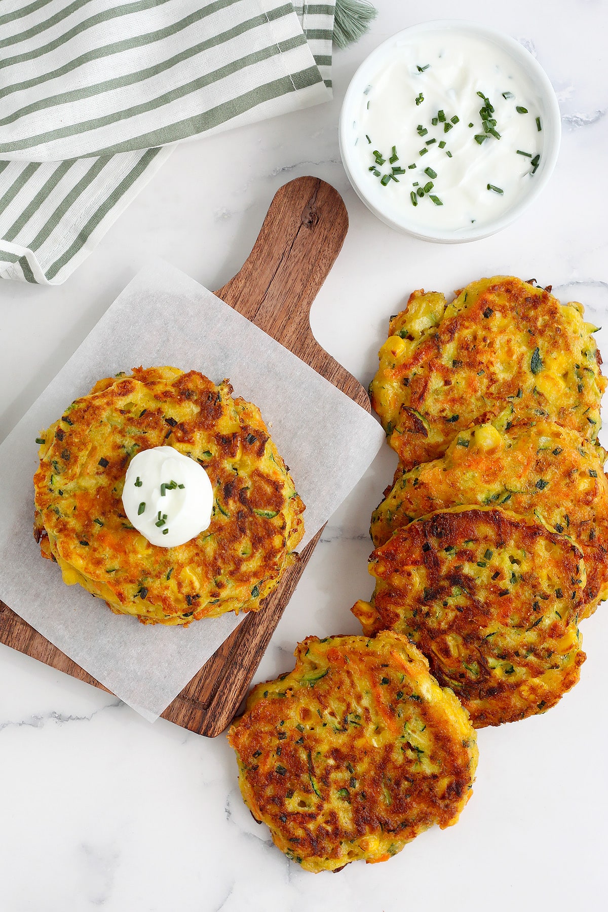 An overhead picture of zucchini pancakes spread out on a marble countertop with sour cream dip and a green striped linen in the background.