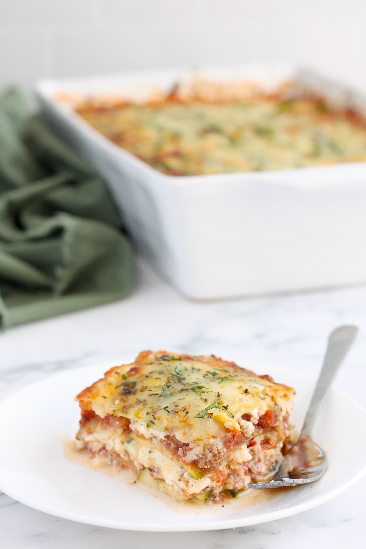 A slice of zucchini lasagna on a white serving plate topped with fresh basil with a fork.