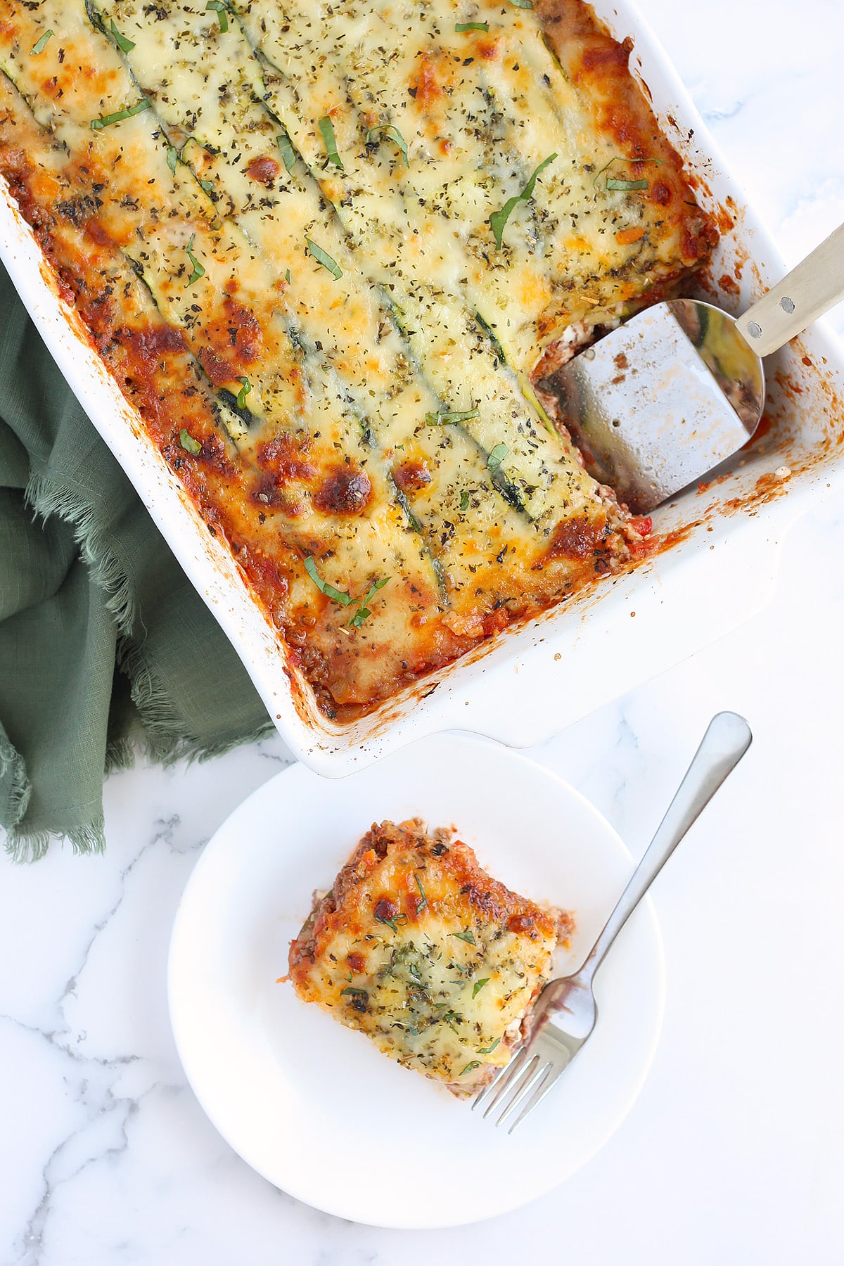 An overhead shot of zucchini lasagna with a slice removed on a white serving plate with a green linen in the background.