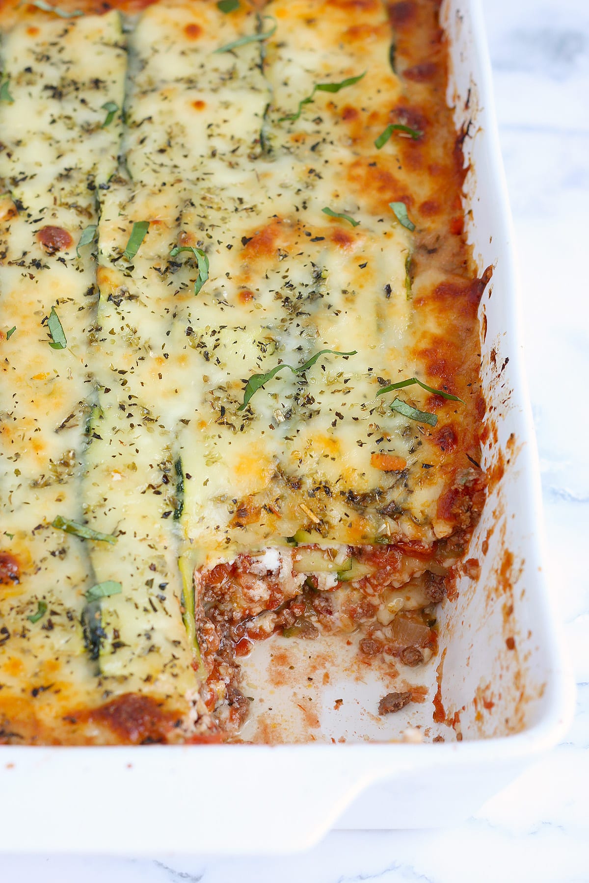A closer up shot of zucchini lasagna with beef in a white ceramic baking dish.