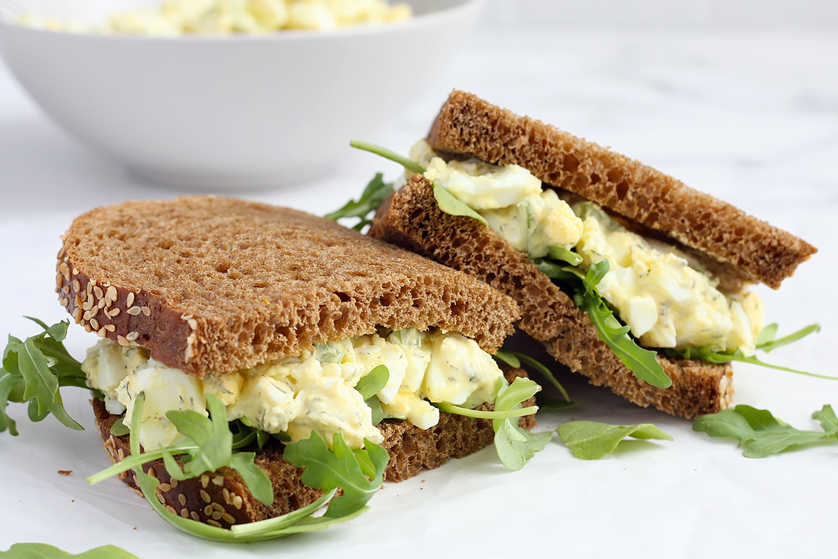 Homemade egg salad sandwiches on sweet brown bread with a bowl of egg salad in the background.