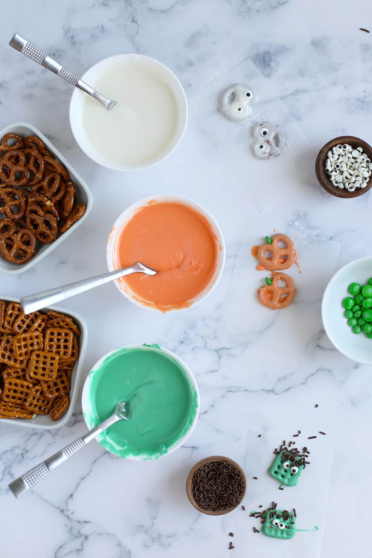 Process photo showing bowls of colored melted chocolate, pretzels, and candies.