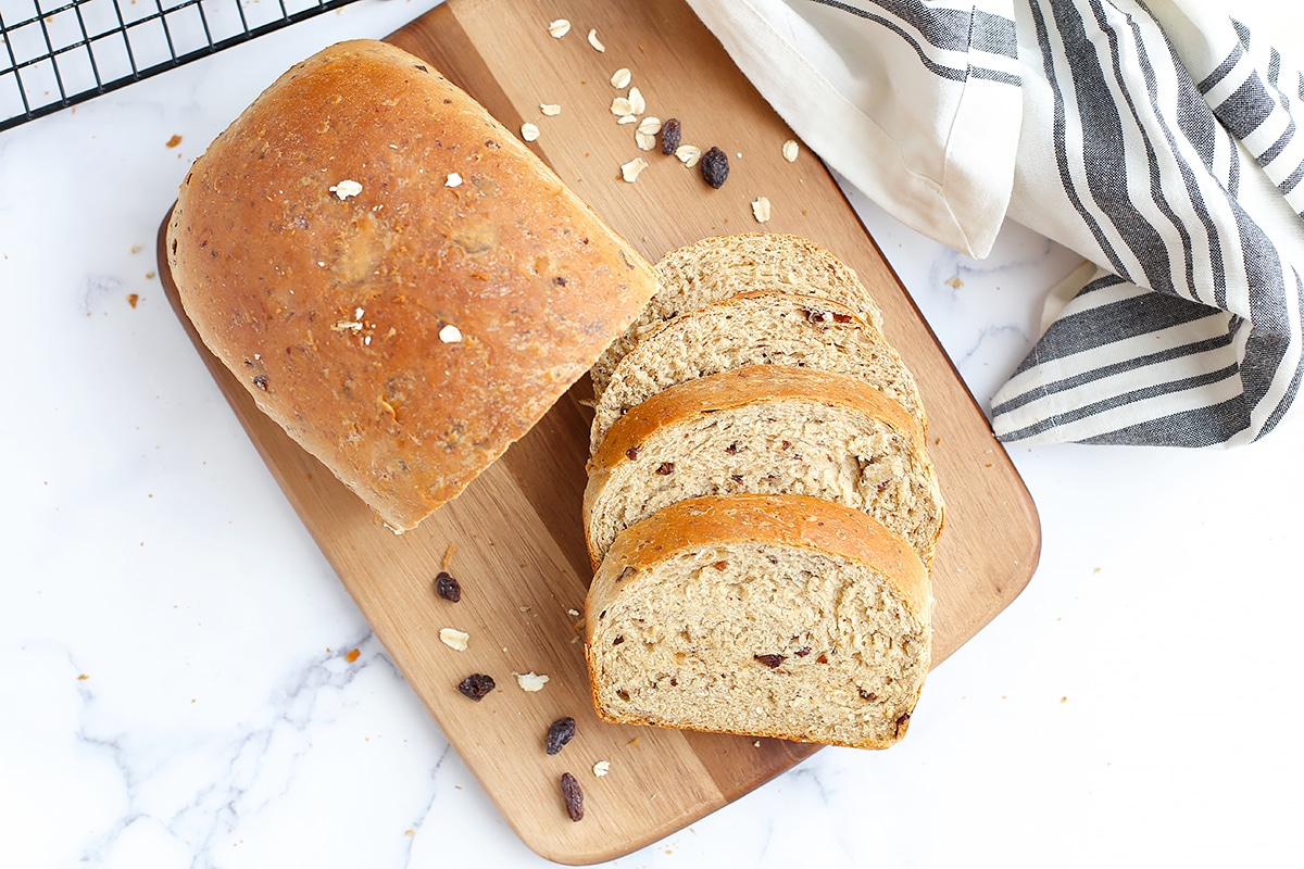 A shot of homemade bread with oats and raisins on a wooden board with a striped linen in the background.