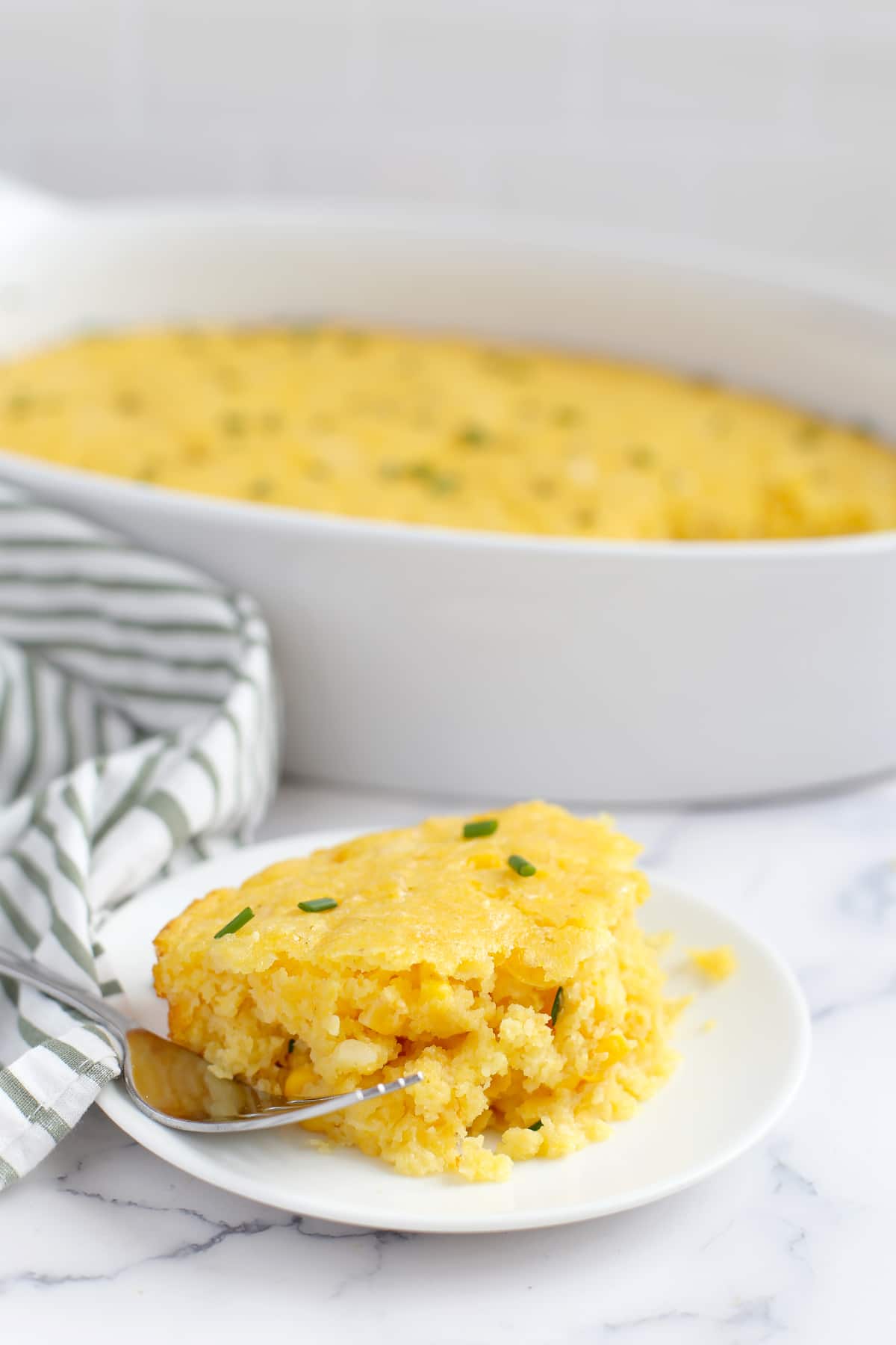 A scoop of cornbread casserole topped with minced chives on a small white serving plate.