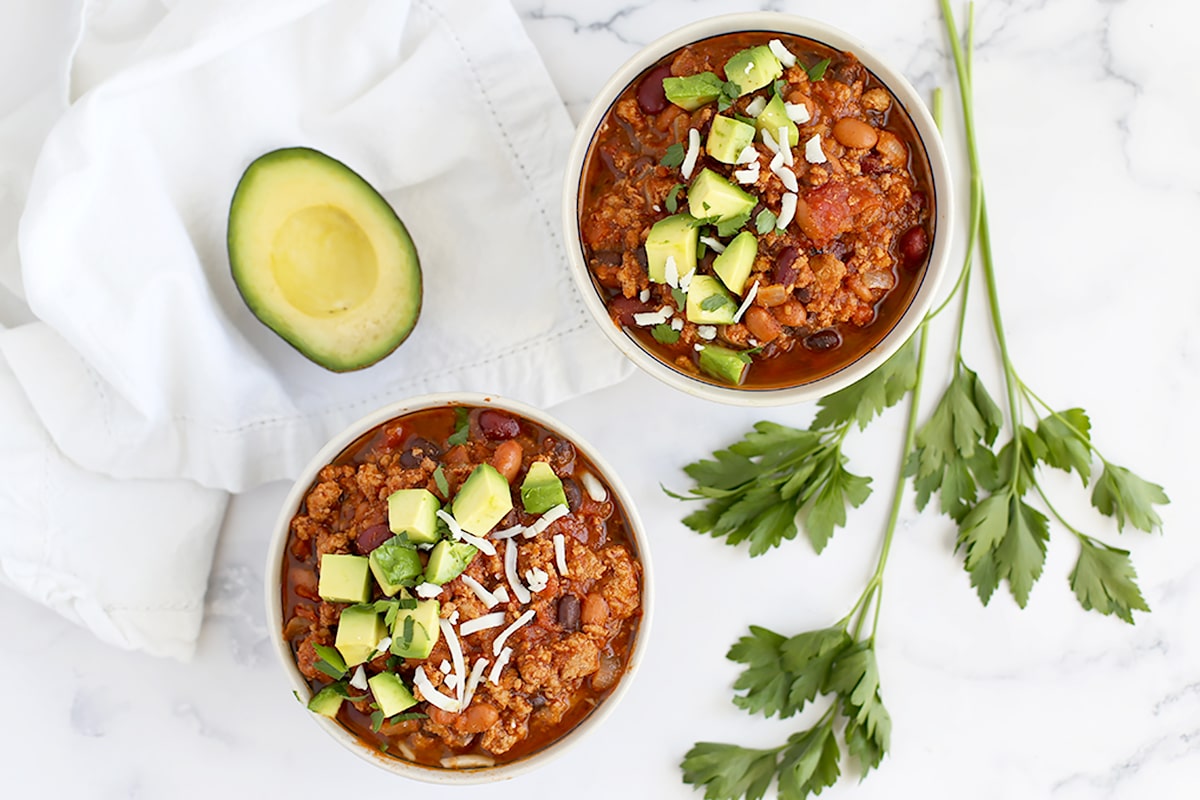 Two bowls of turkey chili with a white linen and half an avocado in the background.