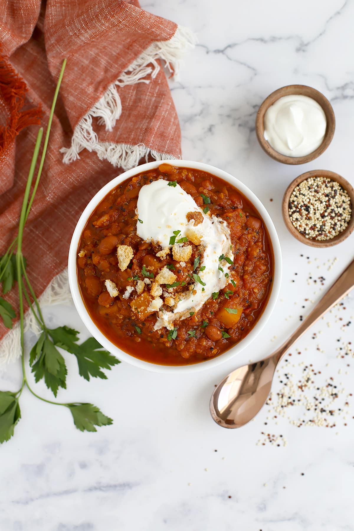 An overhead shot of a bowl of pumpkin chili in a white serving bowl with a sprig of parsley and an orange linen in the background.