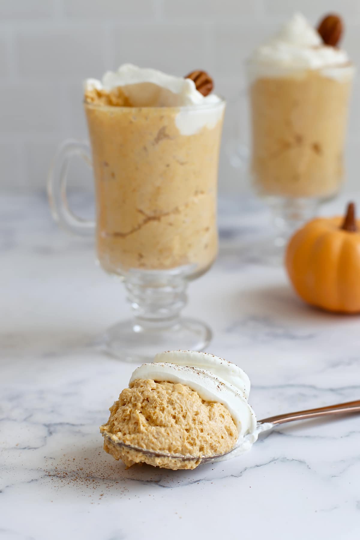 A big spoonful of homemade pumpkin mousse in front of a glass serving jar with a mini pumpkin in the background.