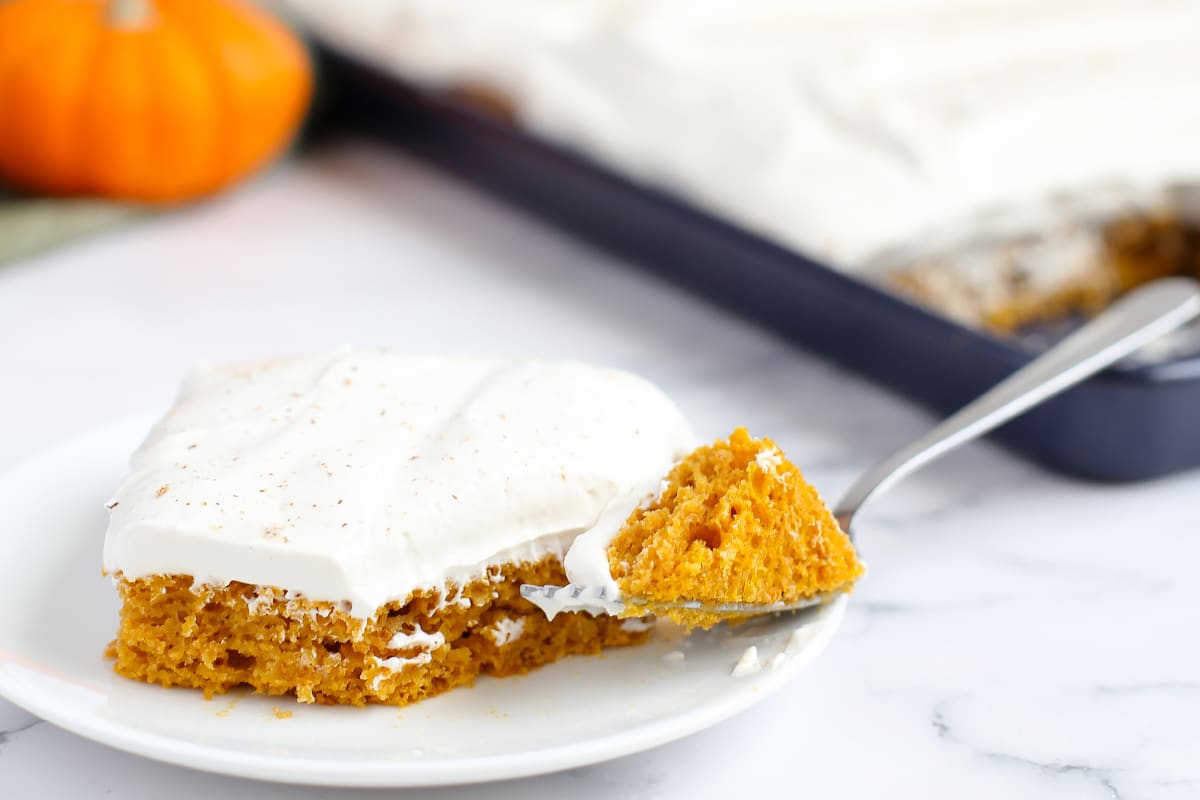 A close up picture of a slice of pumpkin cake with fluffy icing on a serving plate with a fork.