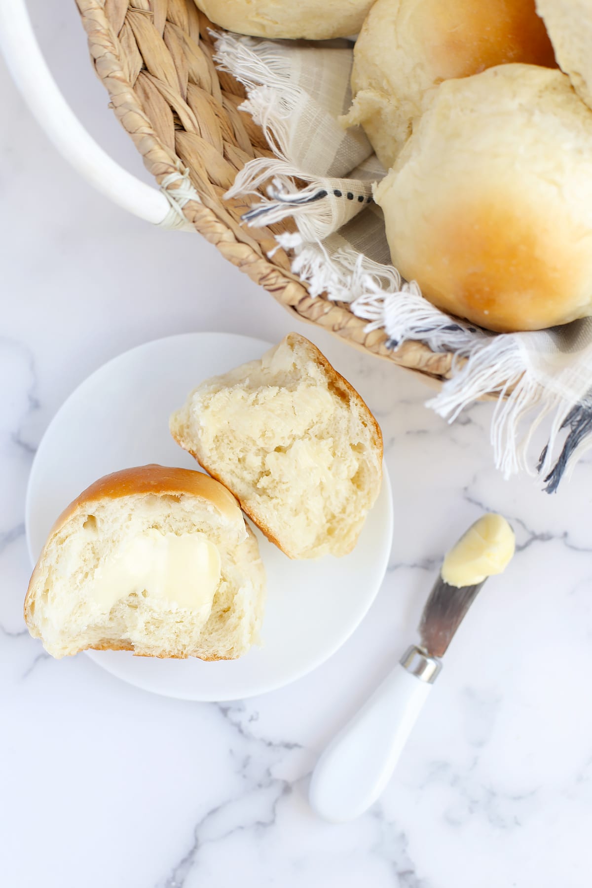 A fluffy dinner roll topped with softened butter on a white serving plate next to a basket of rolls with a checkered linen.