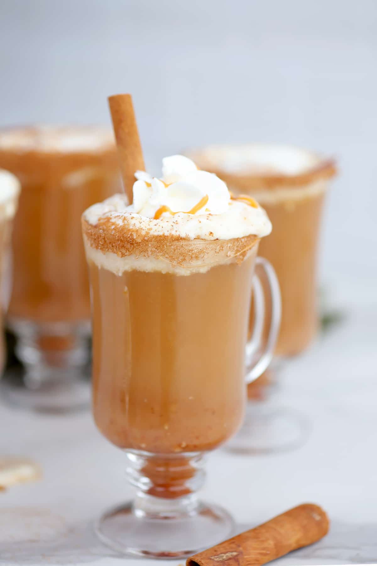 A glass mug filled with caramel apple cider, topped with whipped cream, a caramel drizzle, and a cinnamon stick.