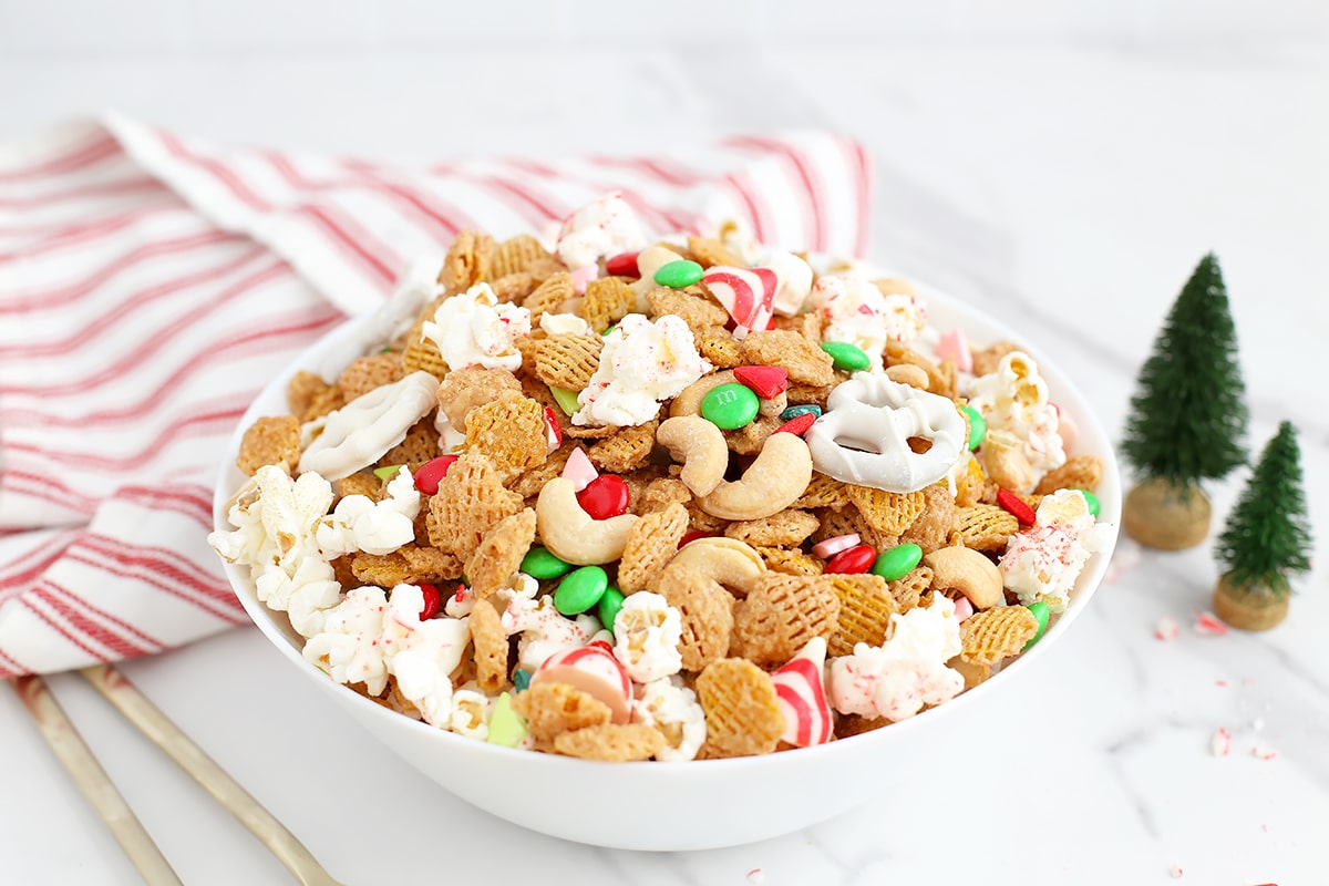 A bowl filled with ،liday snack mix with a red and white ،ed linen and mini christmas trees in the background.