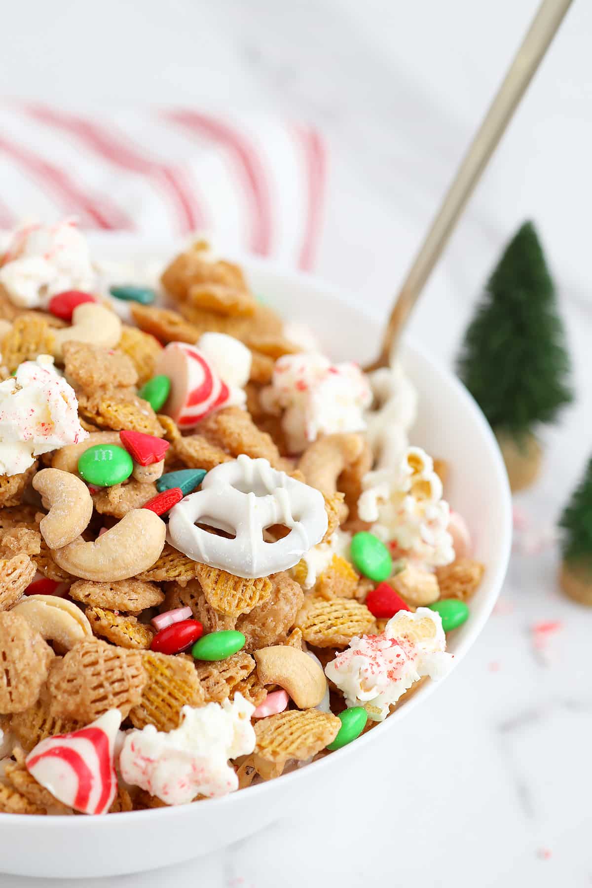 Chocolate covered pretzels, cashews, chex mix, popcorn, and candies tossed together in a white serving bowl with a large spoon. 