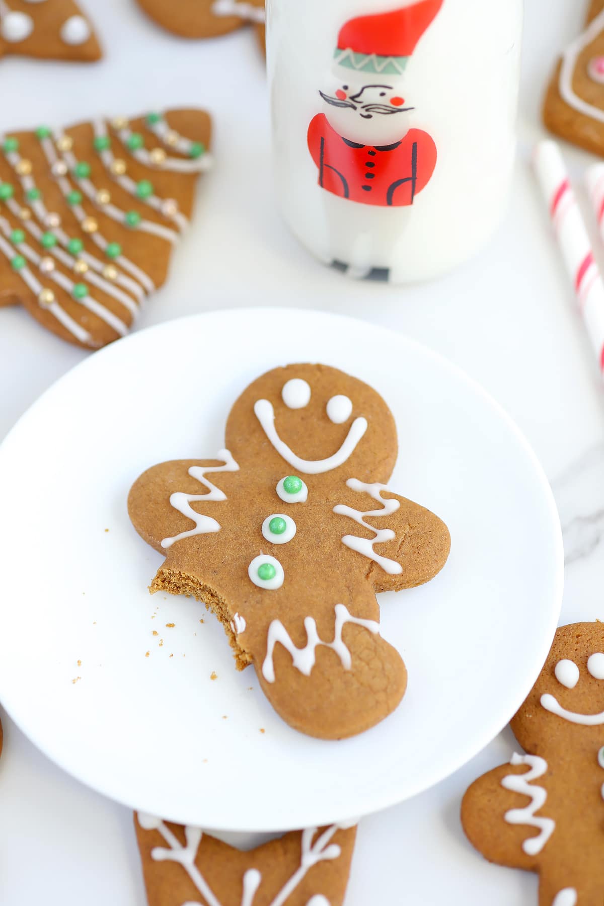 A gingerbread man cookie with a bite taken out on a small white serving plate with a glass of milk in the background.