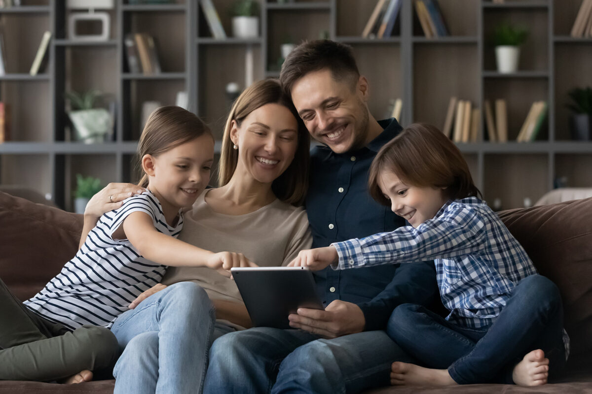 Family of mother, father, and two children sitting on the couch, smiling, all participating in a family meeting using a tablet