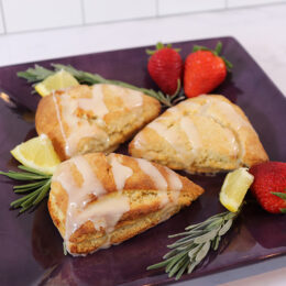Plate of three lavender honey scones drizzled with icing on a black square plate, surrounded by a few lavender sprigs, lemon wedges, and strawberries