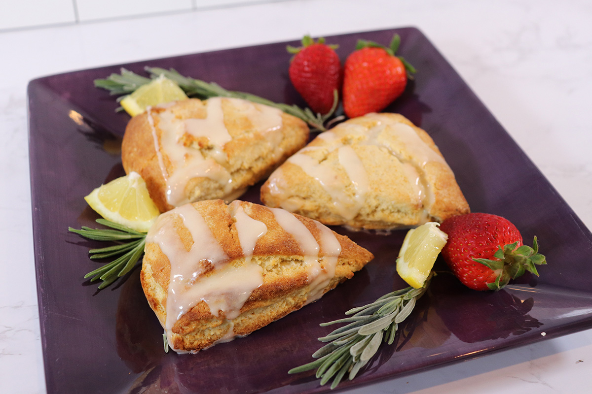 Lavender Honey Scones First Image Plate of three lavender honey scones drizzled with icing on a black square plate, surrounded by a few lavender sprigs, lemon wedges, and strawberries