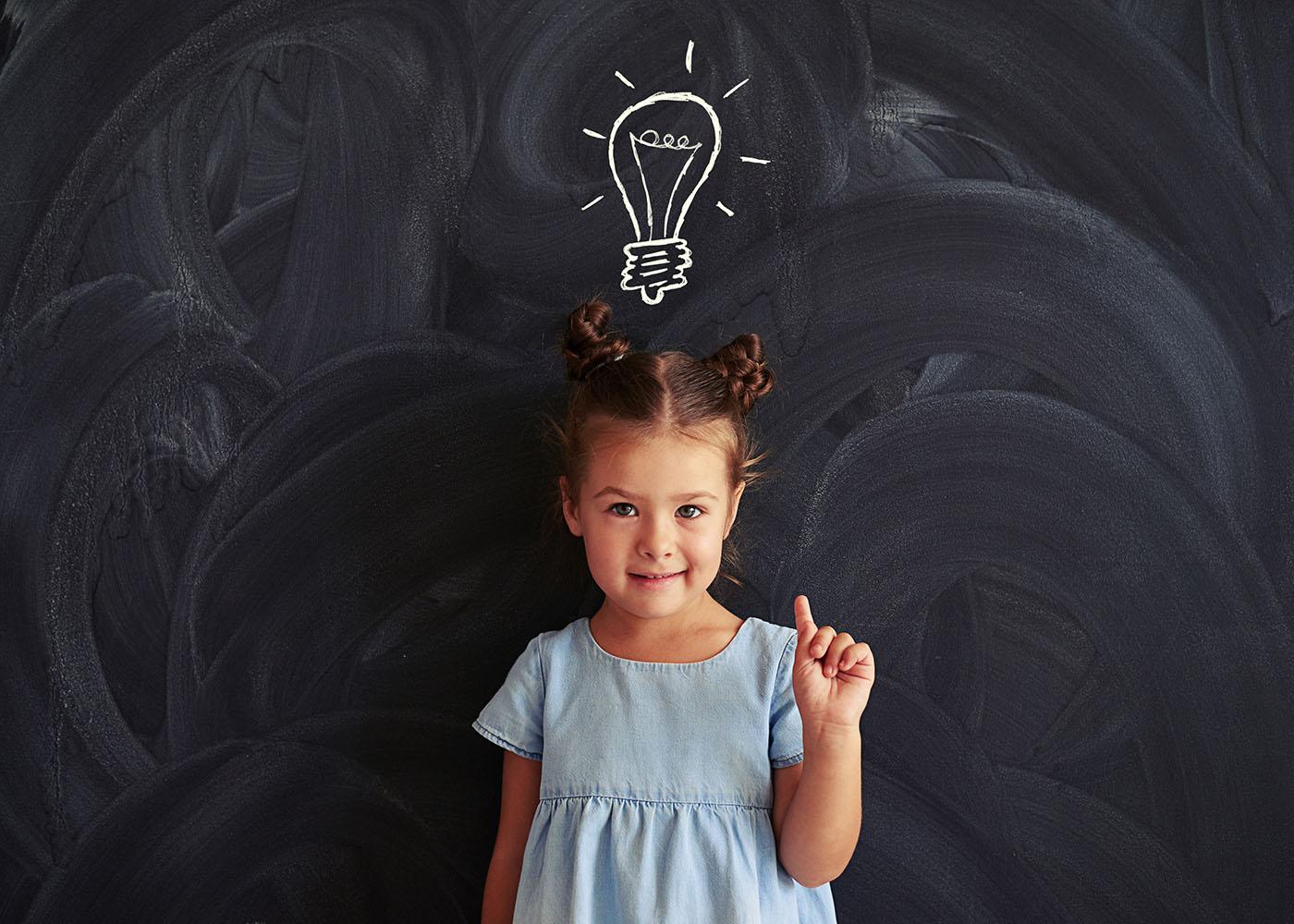 A little brown-haired girl wearing a blue dress holding up one finger standing in front of a chalkboard. On the chalkboard behind her and above her head is the chalk drawing of a lightbulb, as if she has a bright idea.