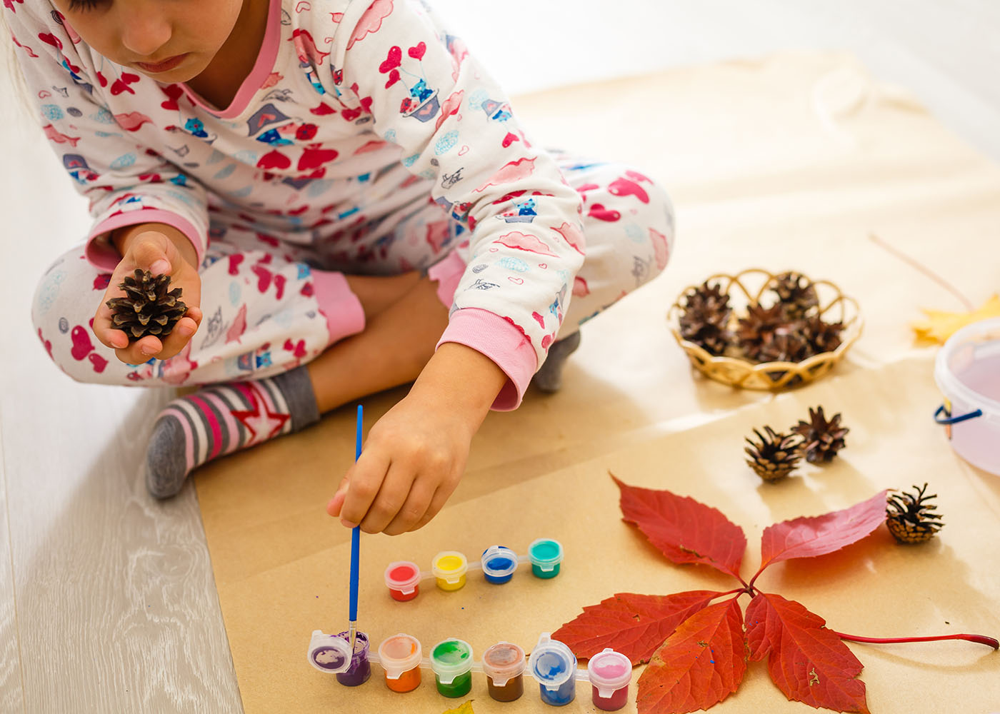 Little girl in colorful pajamas dipping a paintbrush in purple paint to paint a leaf she has in her other hand