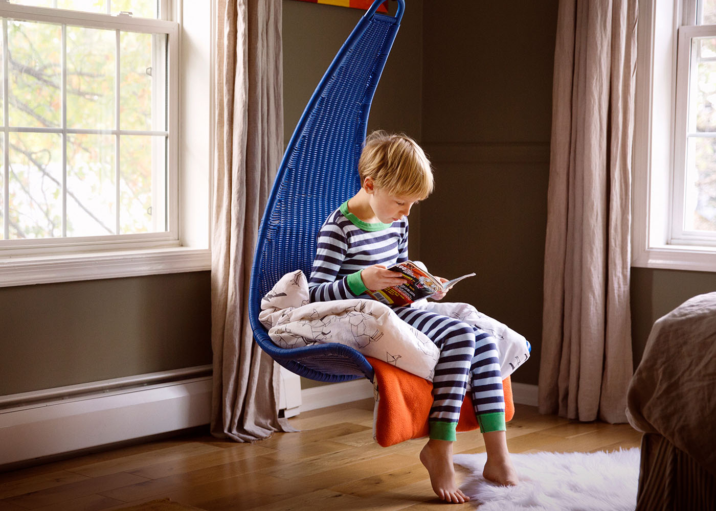 Young boy sitting in blue hanging chair in the corner of a room wearing pajamas and reading a book