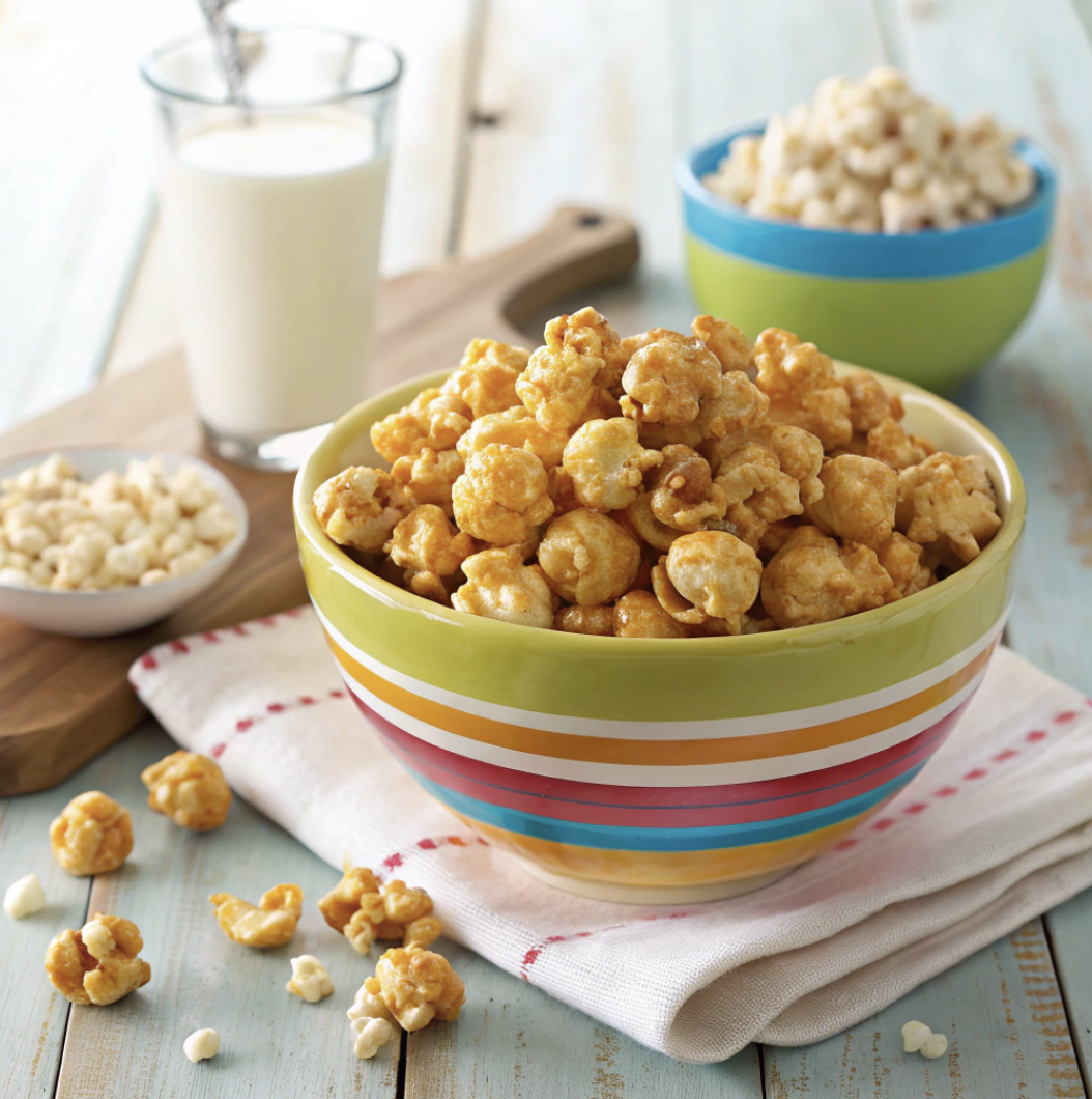 A vibrant bowl of Susan’s Crispy Caramel Corn, filled with golden, glossy caramel-coated popcorn, served in a colorful ceramic bowl on a rustic wooden table. A cozy, family-friendly setting with a checkered napkin, a mug of hot cocoa, and scattered popcorn pieces for a nostalgic vibe. Soft natural light highlights the caramel’s shine and popcorn’s texture, with a shallow depth of field and a warm kitchen background. Professional food photography style, inspired by SuperHealthyKids.com